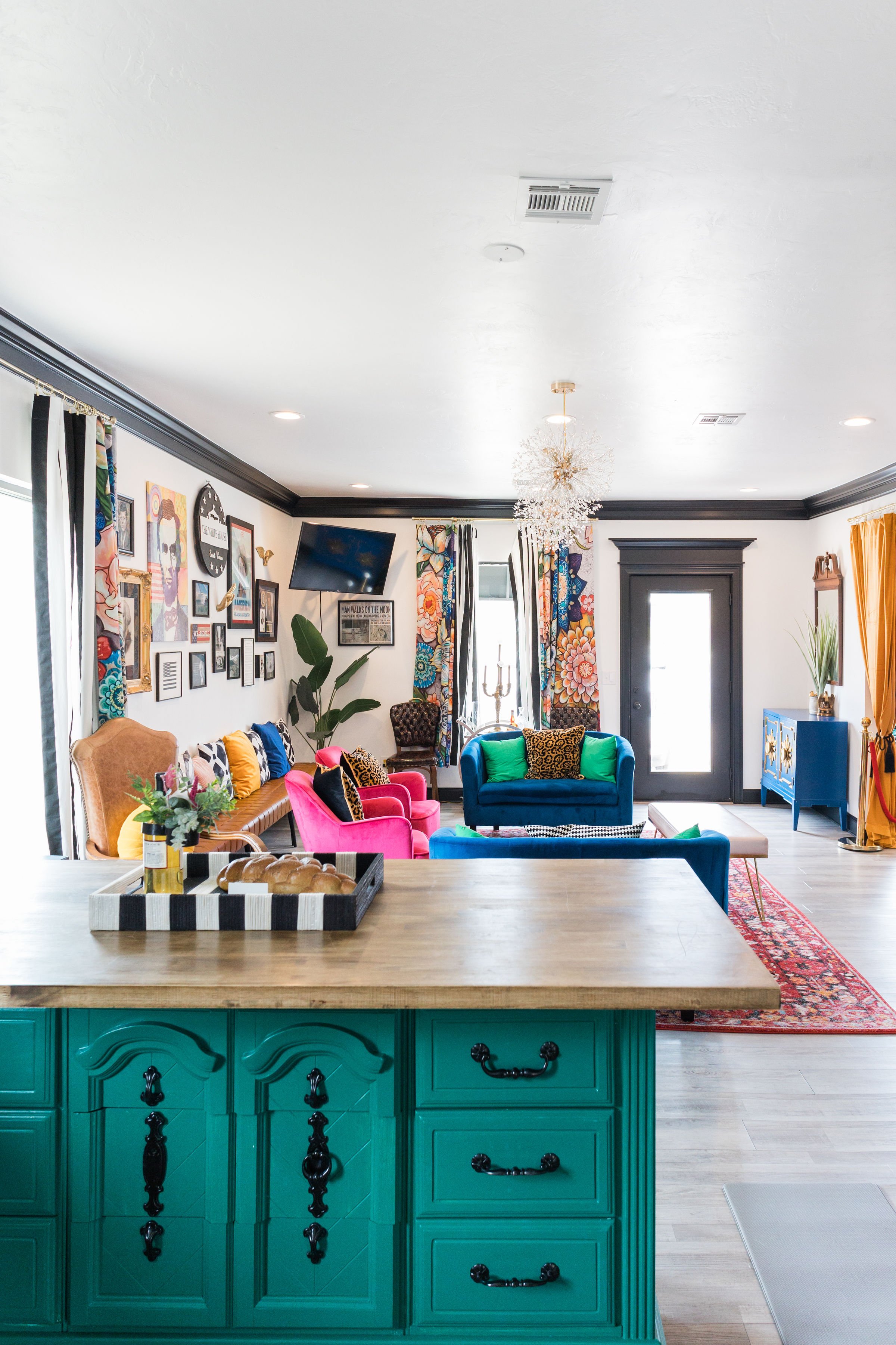 A colorful living room with an open kitchen in the foreground, featuring turquoise cabinets and a wooden countertop with a tray of bread. The living room has a variety of vibrant furniture, including a yellow and tan sofa, pink chairs, a dark blue co