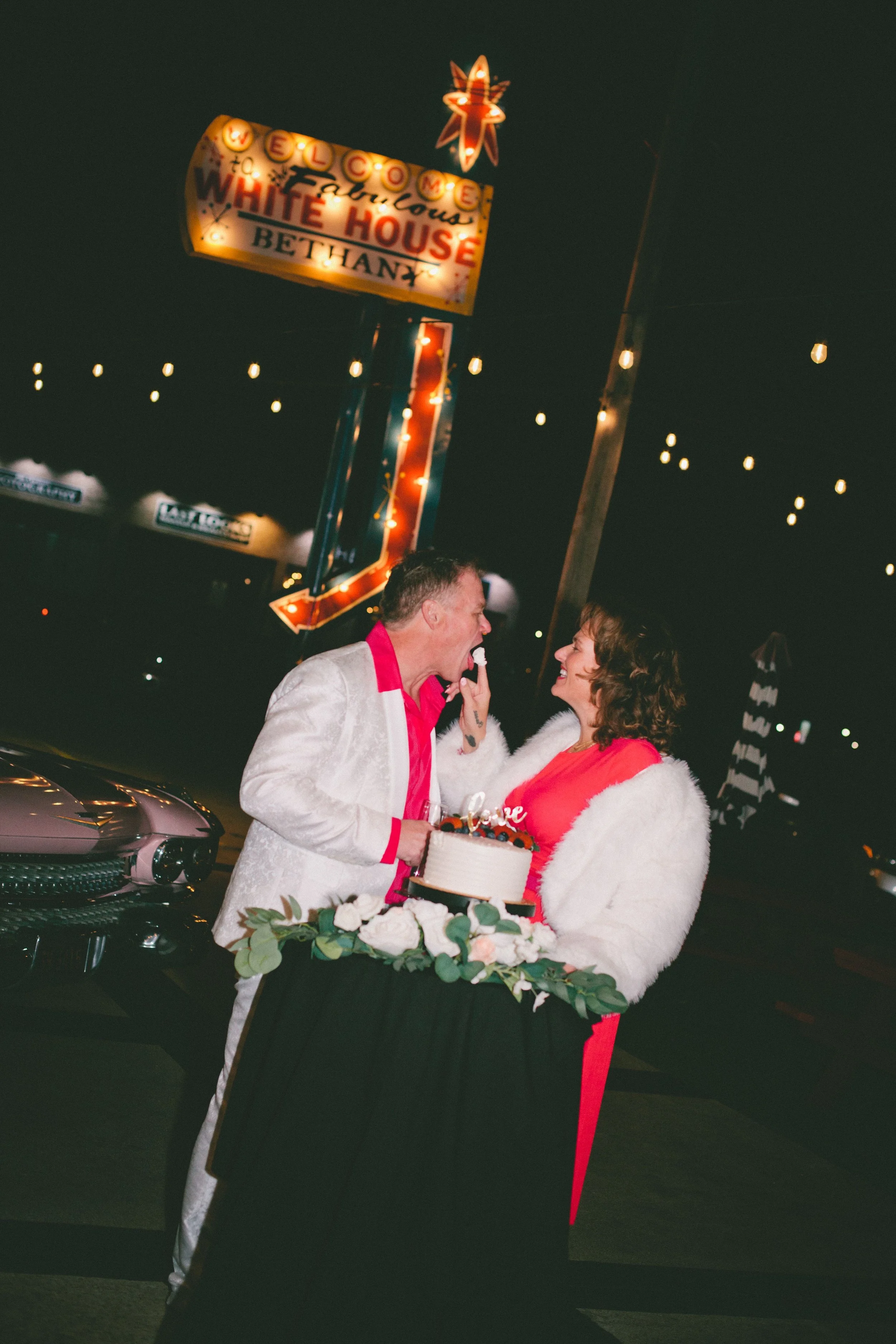 A couple celebrating at night, holding a birthday cake, with a neon sign in the background that reads 'Welcome Fabulous White House Bethany' and string lights overhead.