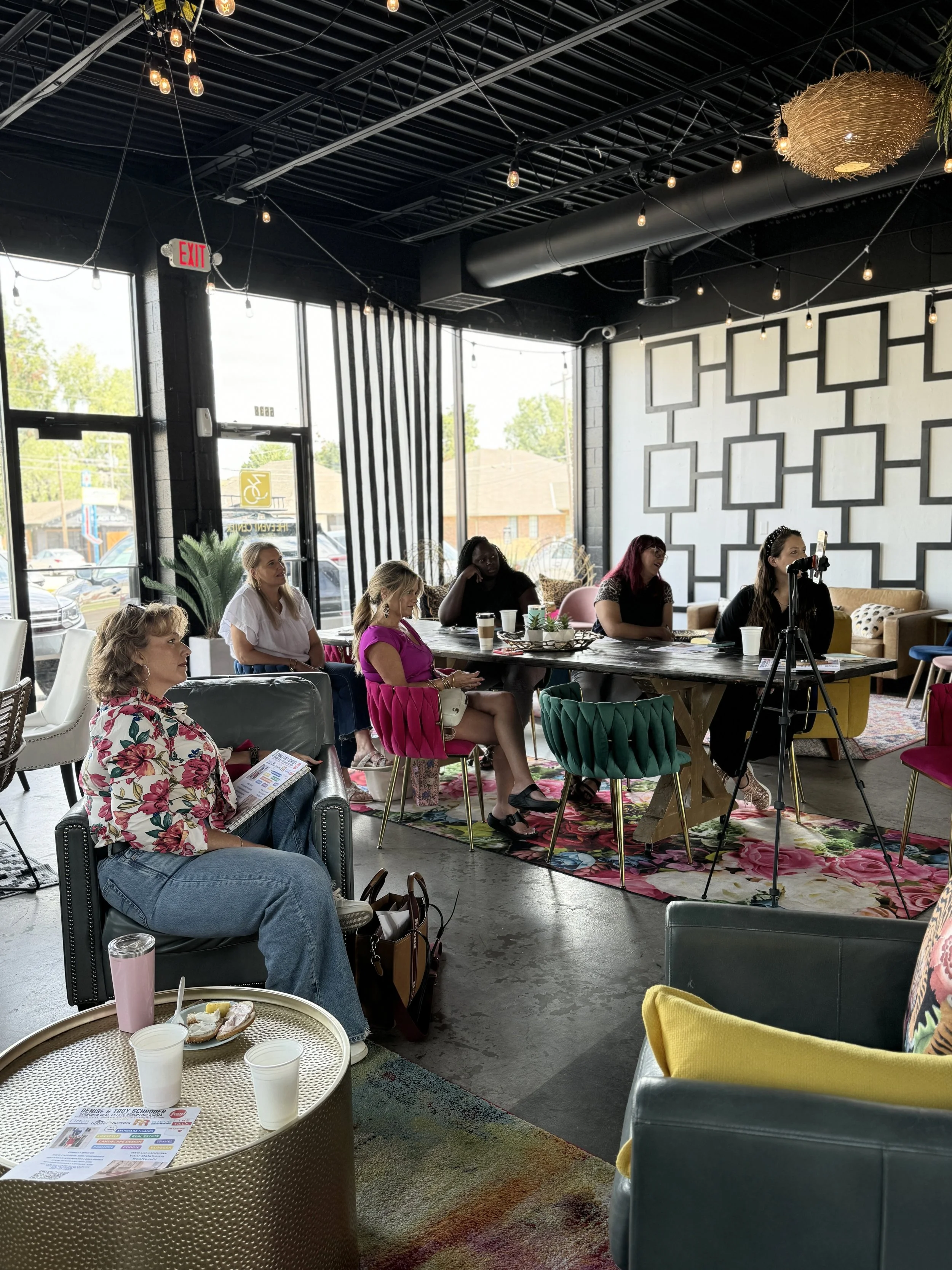 Women attending a meeting or workshop in a brightly decorated coffee shop with modern design, floral rugs, and large windows.