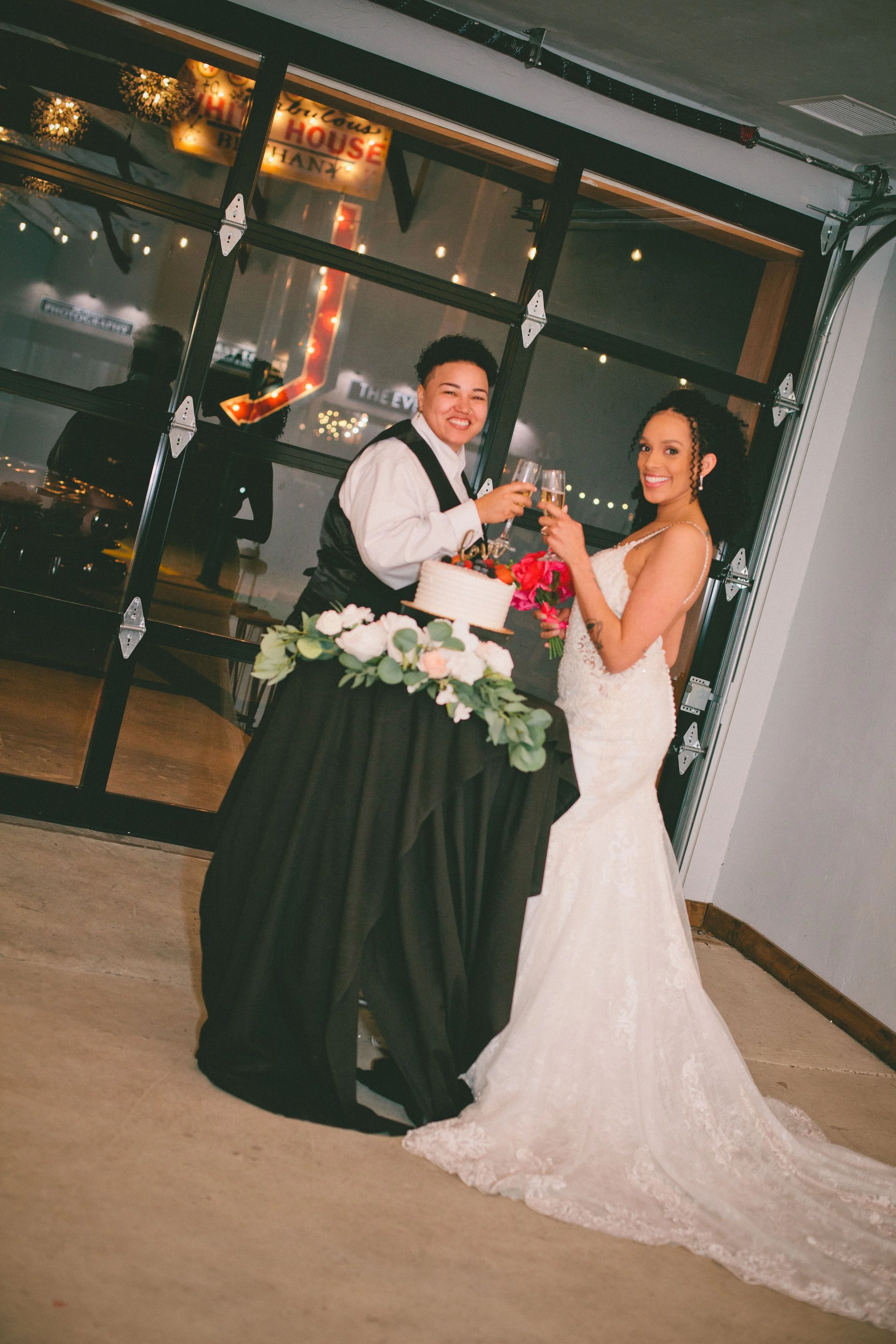 Two women, one in a wedding dress and the other in a tuxedo, are celebrating with champagne at a wedding reception, standing next to a table with a cake and floral decorations.