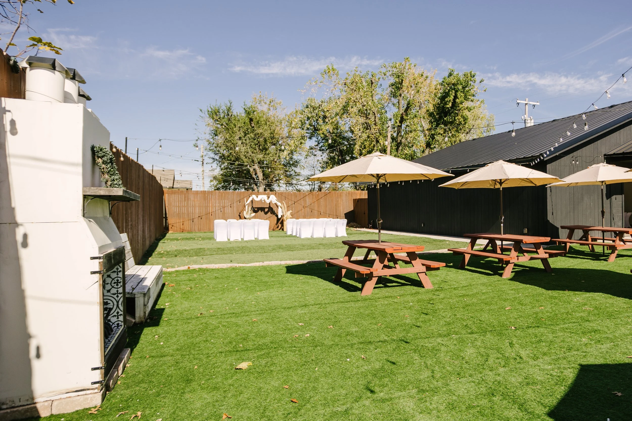Backyard with green grass, outdoor tables with umbrellas, white chairs, and a decorated arch for an event. String lights above and trees in the background.