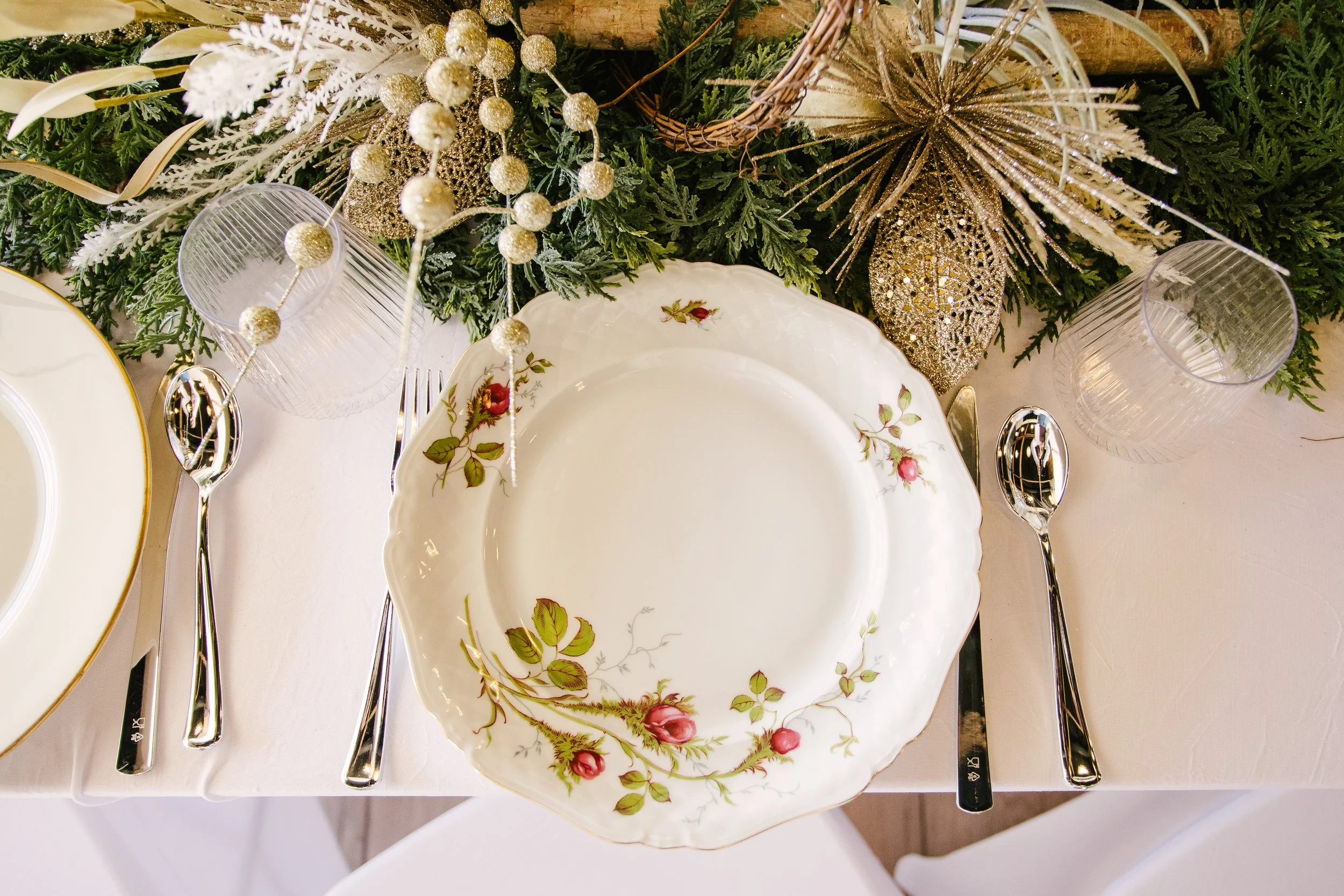 Elegant holiday table setting with a floral-patterned china plate, silver utensils, glassware, and festive decorations including gold ornaments, greenery, and ribbon.