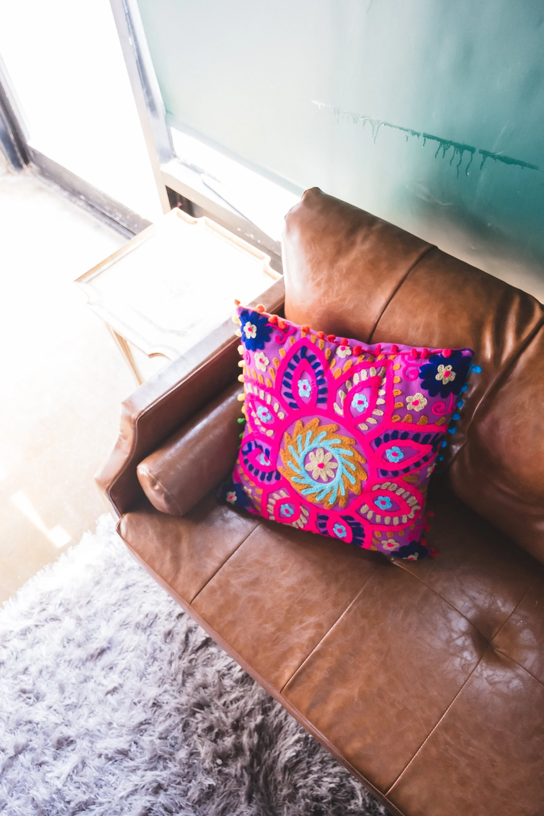 A leather sofa with a colorful embroidered cushion placed on it, next to a small table near a bright window.