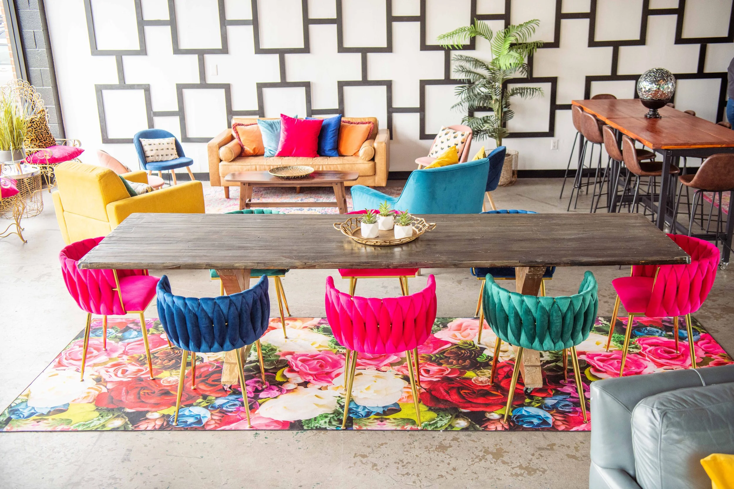 Colorful modern living room with a wooden dining table, pink and teal velvet chairs, a floral area rug, and a cozy seating area with a beige sofa, vibrant pillows, and a variety of colorful chairs.