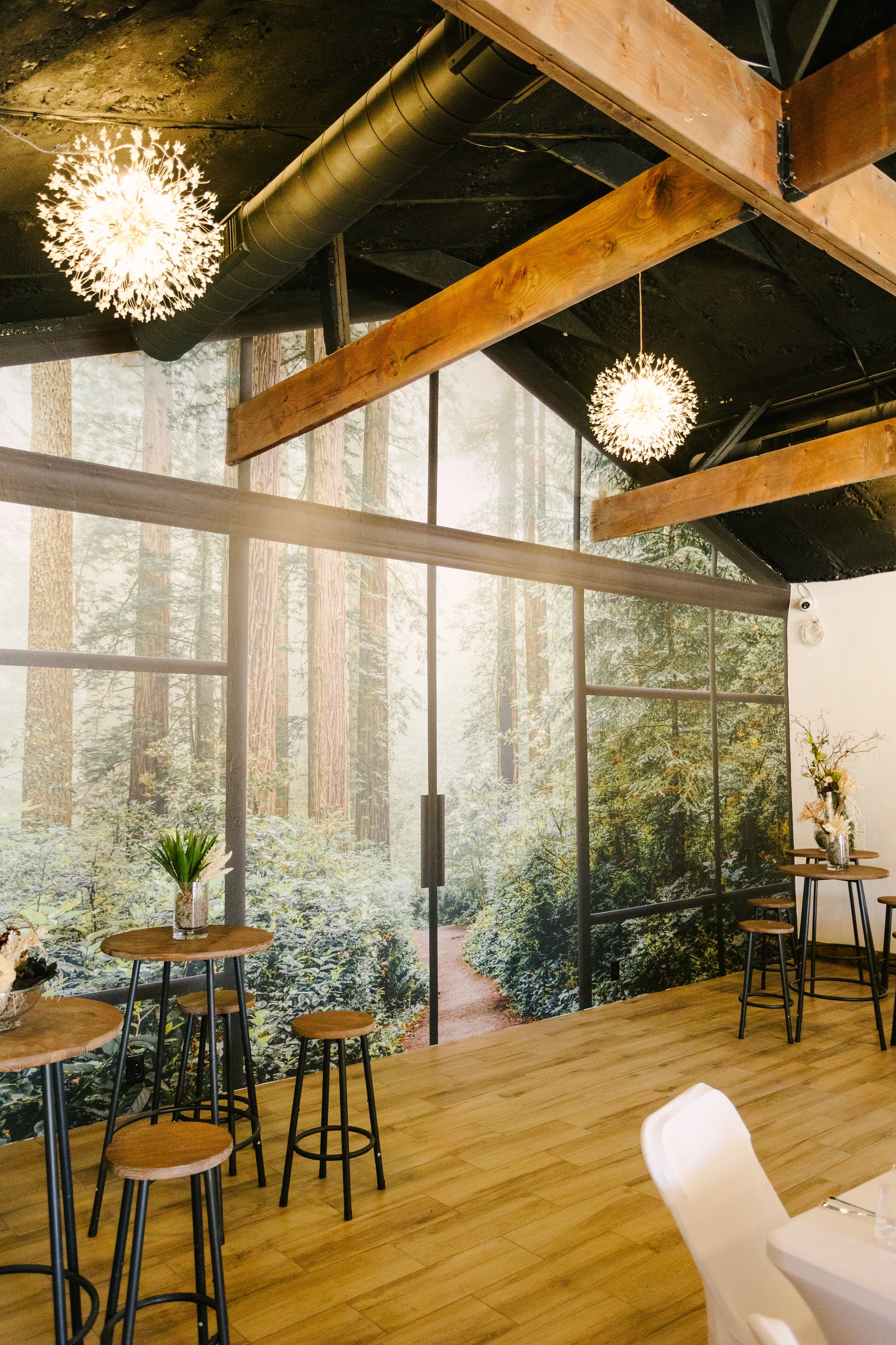 Indoor space with large forest-themed mural wall, wooden beams, hanging spherical light fixtures, and small round tables with flower arrangements.