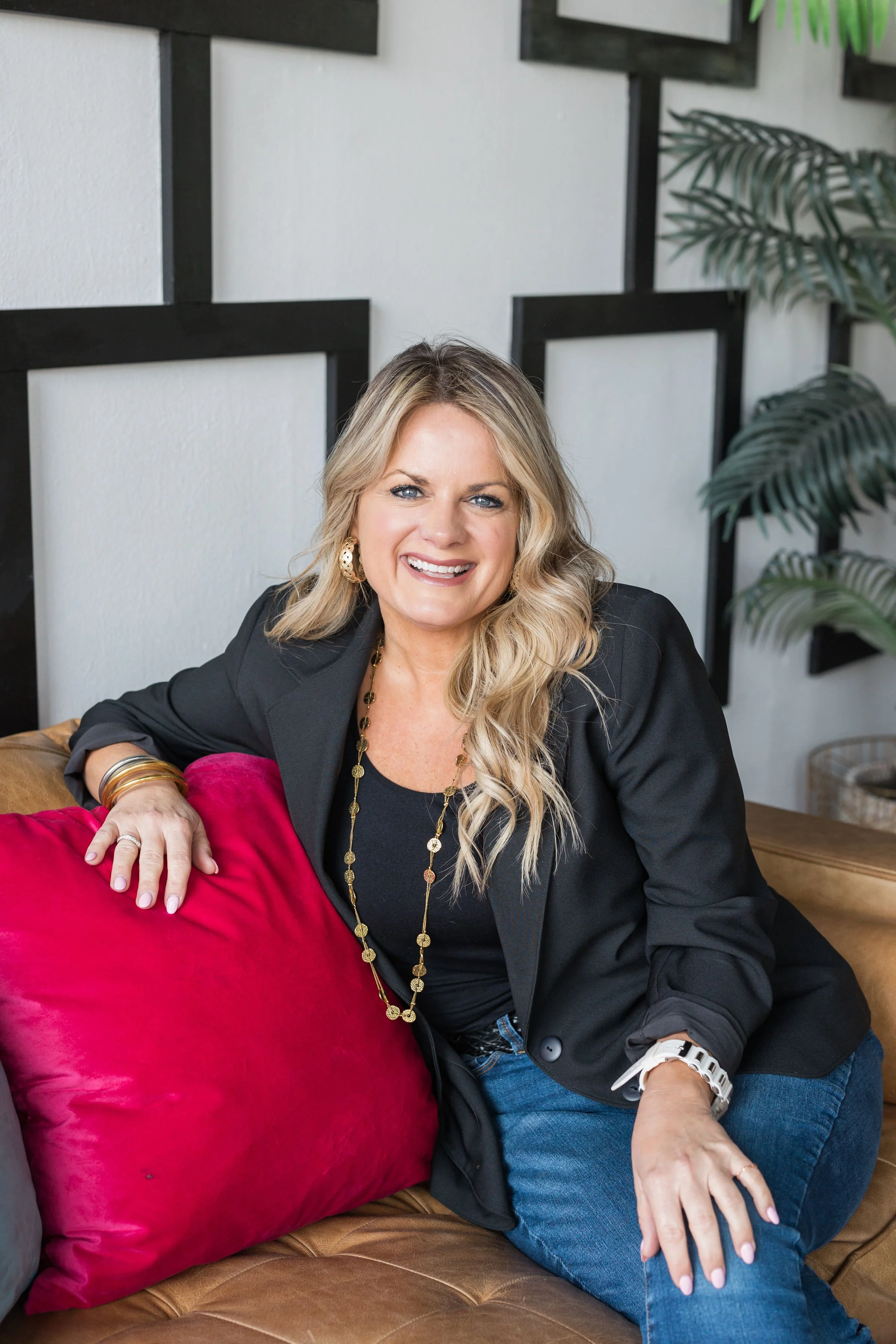 A smiling woman with blond hair, wearing a black blazer and jeans, sitting on a brown leather couch with a red pillow. She is accessorized with gold jewelry and a white watch, in a modern setting with plants and geometric wall decor.