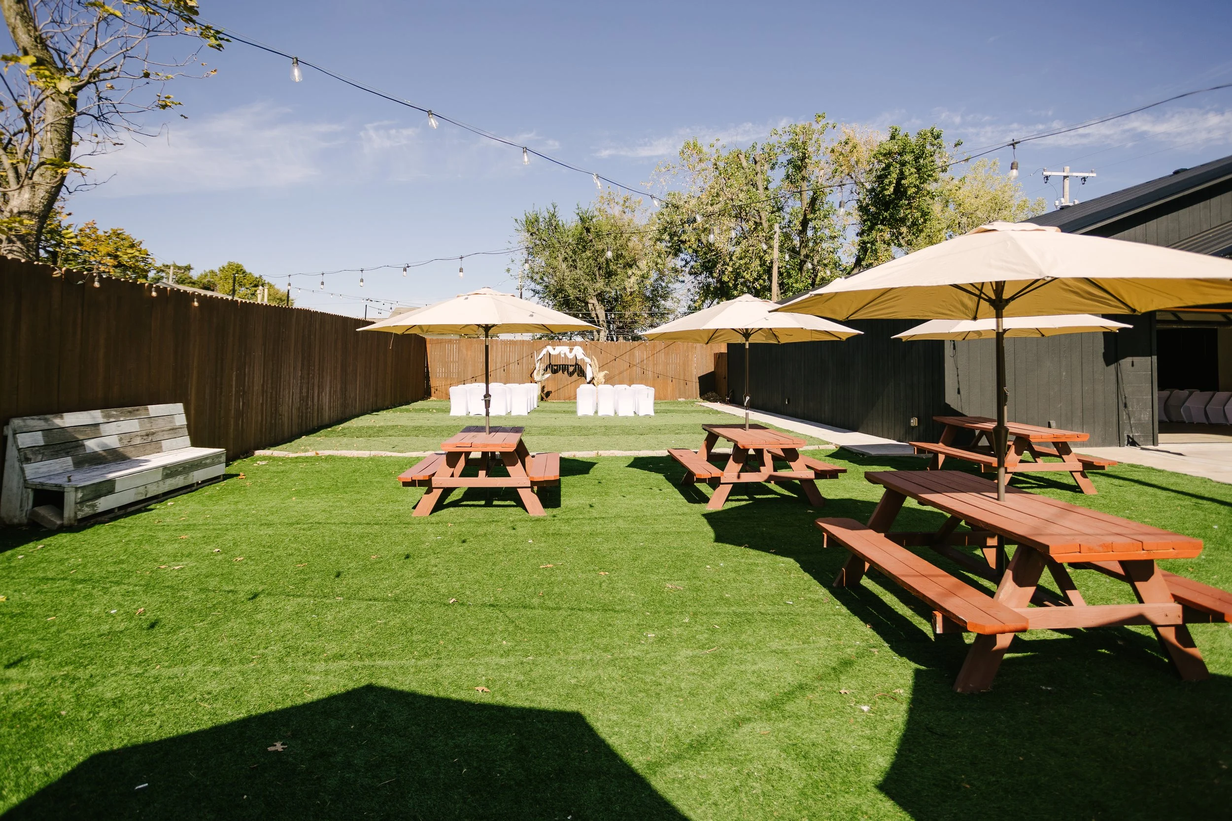 Outdoor yard with wooden picnic tables under umbrellas, string lights, a wooden fence, and a white decorated arch at the back on a sunny day.