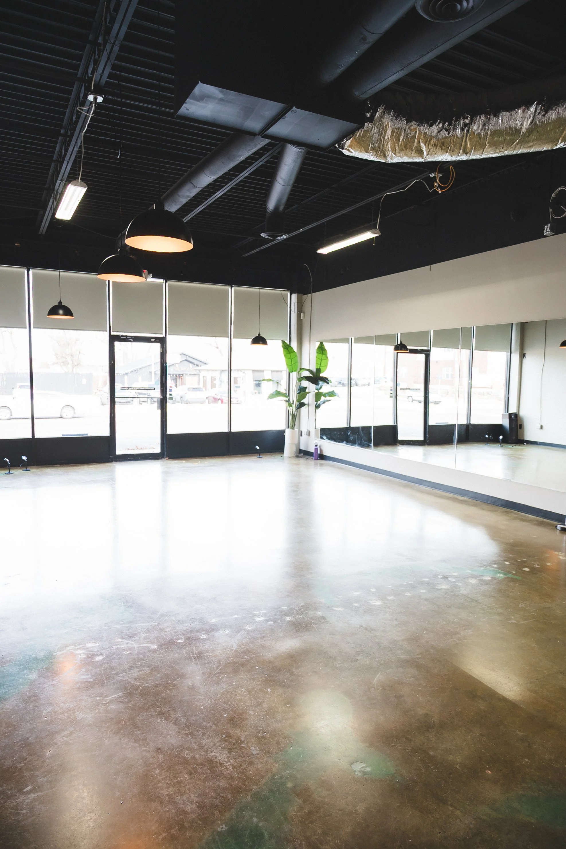 Empty dance studio with large wall mirrors, black ceiling with exposed ductwork, pendant lights, and a potted plant near the windows.