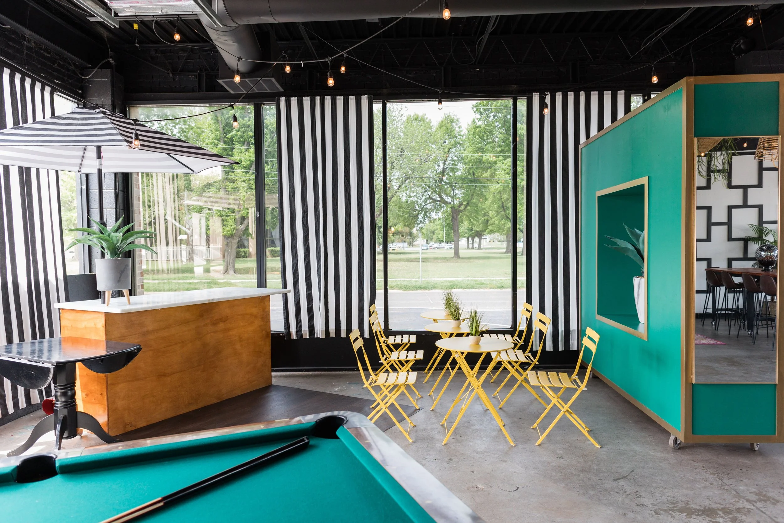 Interior of a modern cafe with yellow chairs, small round white tables with plants, a pool table in the foreground, black and white striped walls, large windows showing a green outdoor area, a teal booth with a cutout, and string lights hanging from 