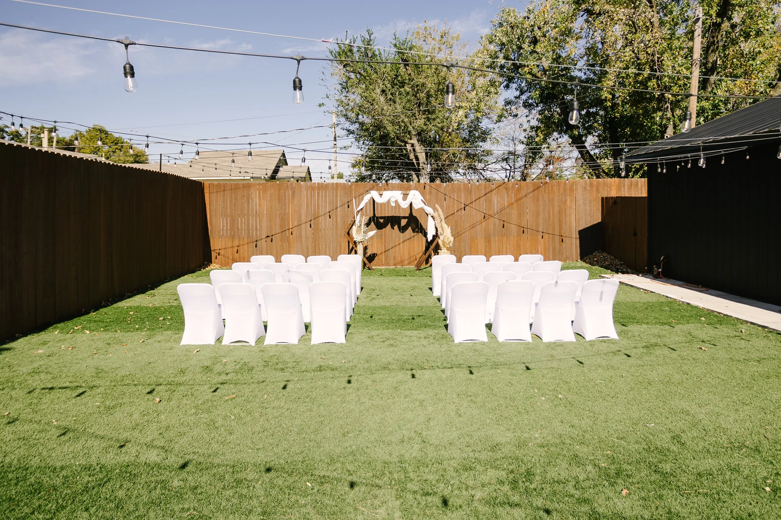 Backyard with white chairs arranged in rows facing a decorated arch, set on a lawn with string lights overhead, wooden fences on sides, and trees in the background, prepared for a wedding or outdoor event.