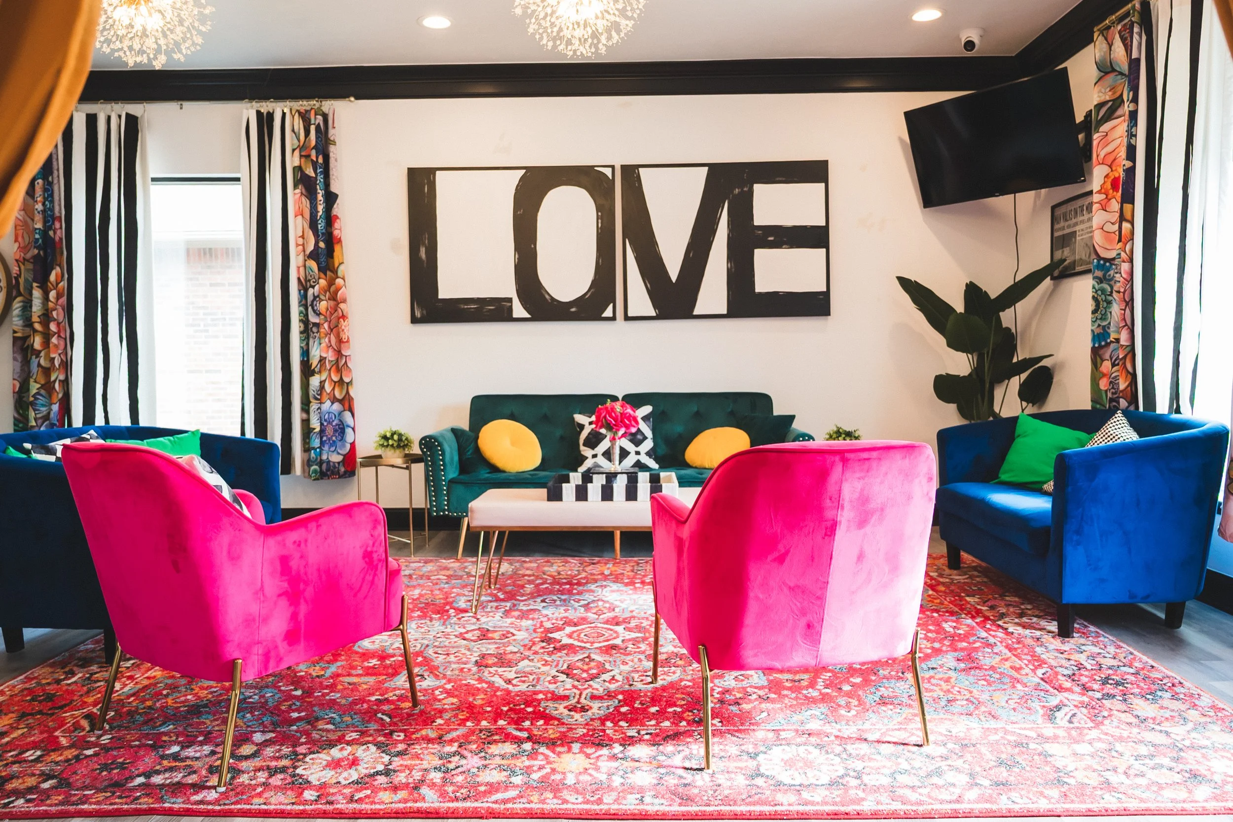 Colorful living room with a green sofa, two pink velvet armchairs, two blue velvet armchairs, a patterned rug, and wall art spelling 'LOVE' in black and white. Features floral curtains, a large plant, and a wall-mounted TV.