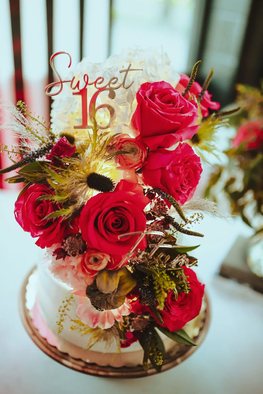 A floral cake decorated with pink and white roses, greenery, and a 'Sweet 16' topper, set against a blurred background.