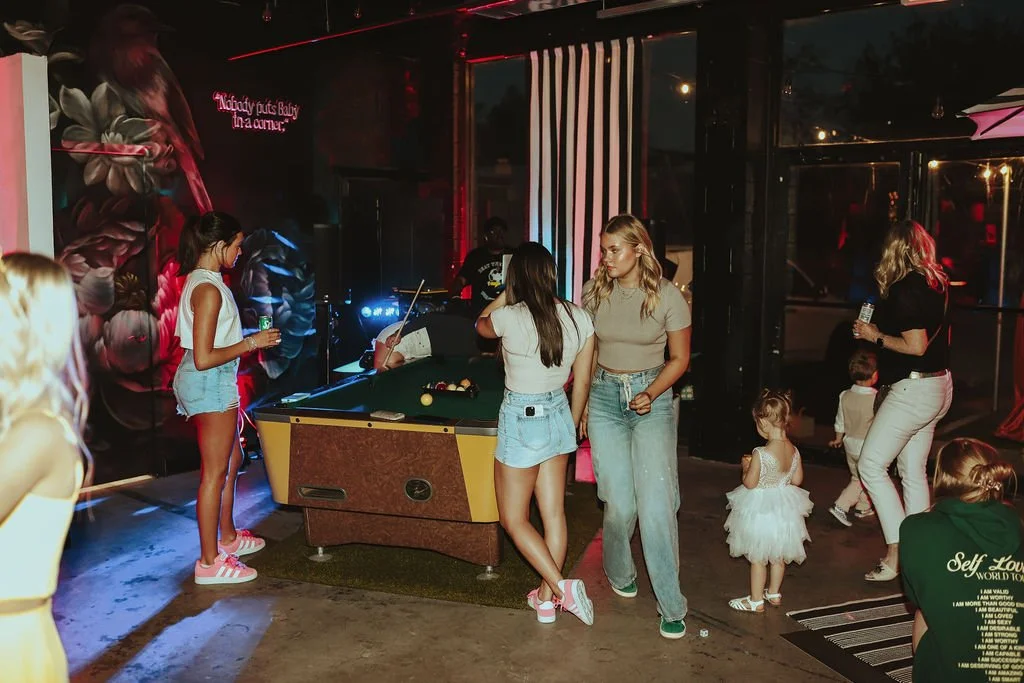 Group of young women and children playing and socializing at a bar or club with a pool table, neon signs, and colorful lighting.