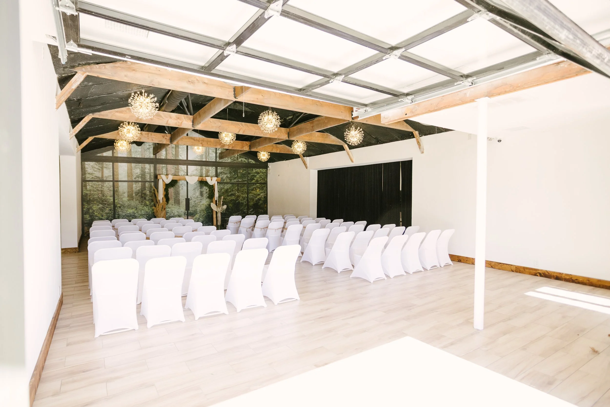 An indoor wedding ceremony setup with white chairs arranged in rows facing an arch decorated with white and gold fabric. The room has wooden beams on the ceiling with decorative light fixtures hanging. The back wall has a large glass window or door s