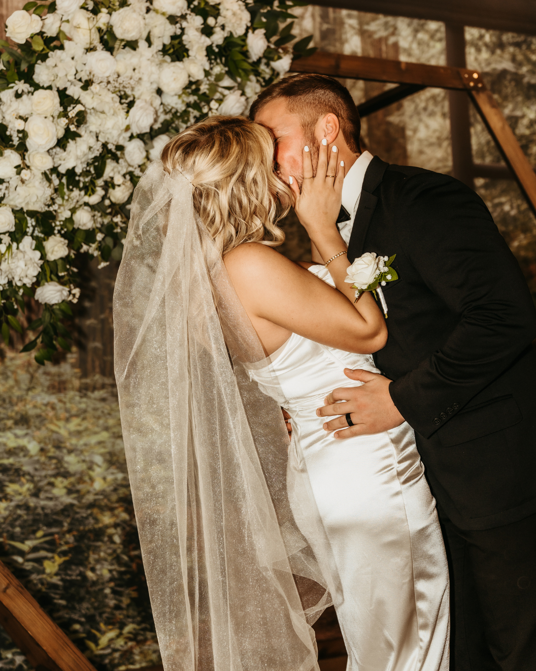 A man and woman kissing in front of an arch on their wedding day. The background has a wall that has a scenic view of a forest that always stays green. 