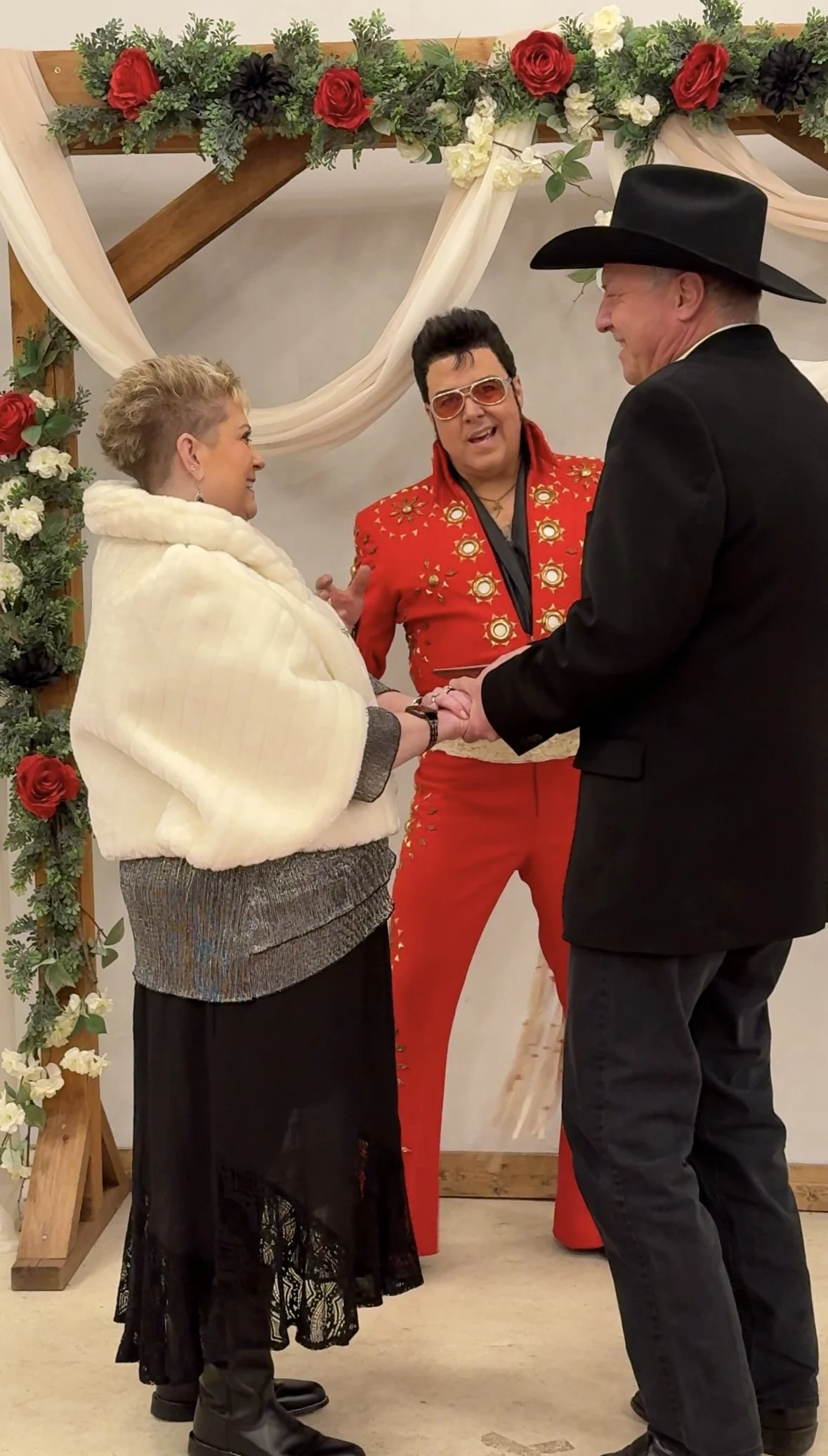 A couple gets married in a rustic indoor wedding ceremony. The bride wears a white cape over a dark dress, and the groom wears a black tuxedo with a cowboy hat. The officiant dressed in a red Elvis Presley-style jumpsuit with sunglasses is conducting
