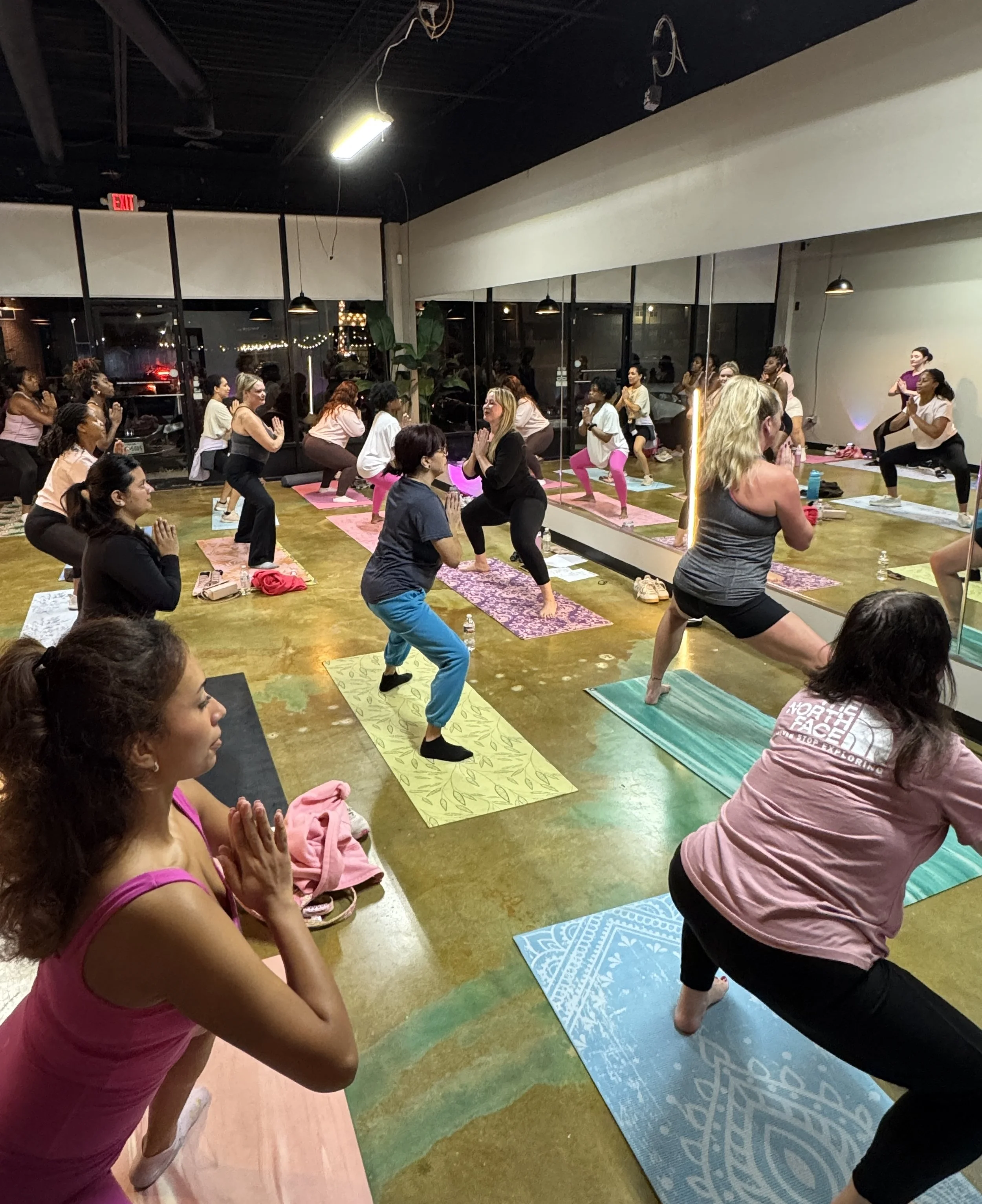 A group of women participating in a yoga or fitness class in a studio with large mirrors and windows at night, practicing yoga poses on colorful mats.