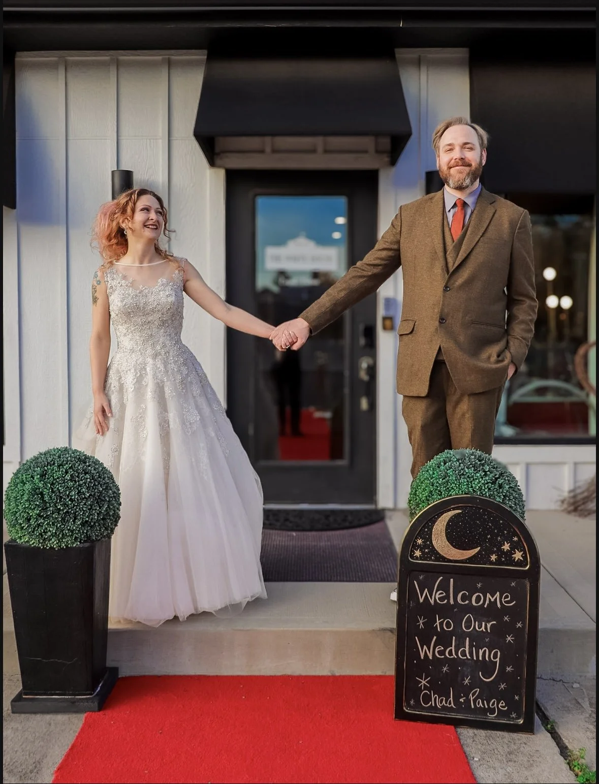 A bride and groom holding hands outside a building, celebrating their wedding. The bride is wearing a white wedding gown and the groom is in a brown suit. A welcome sign with moon and stars drawing reads 'Welcome to Our Wedding Chad & Paige,' placed 