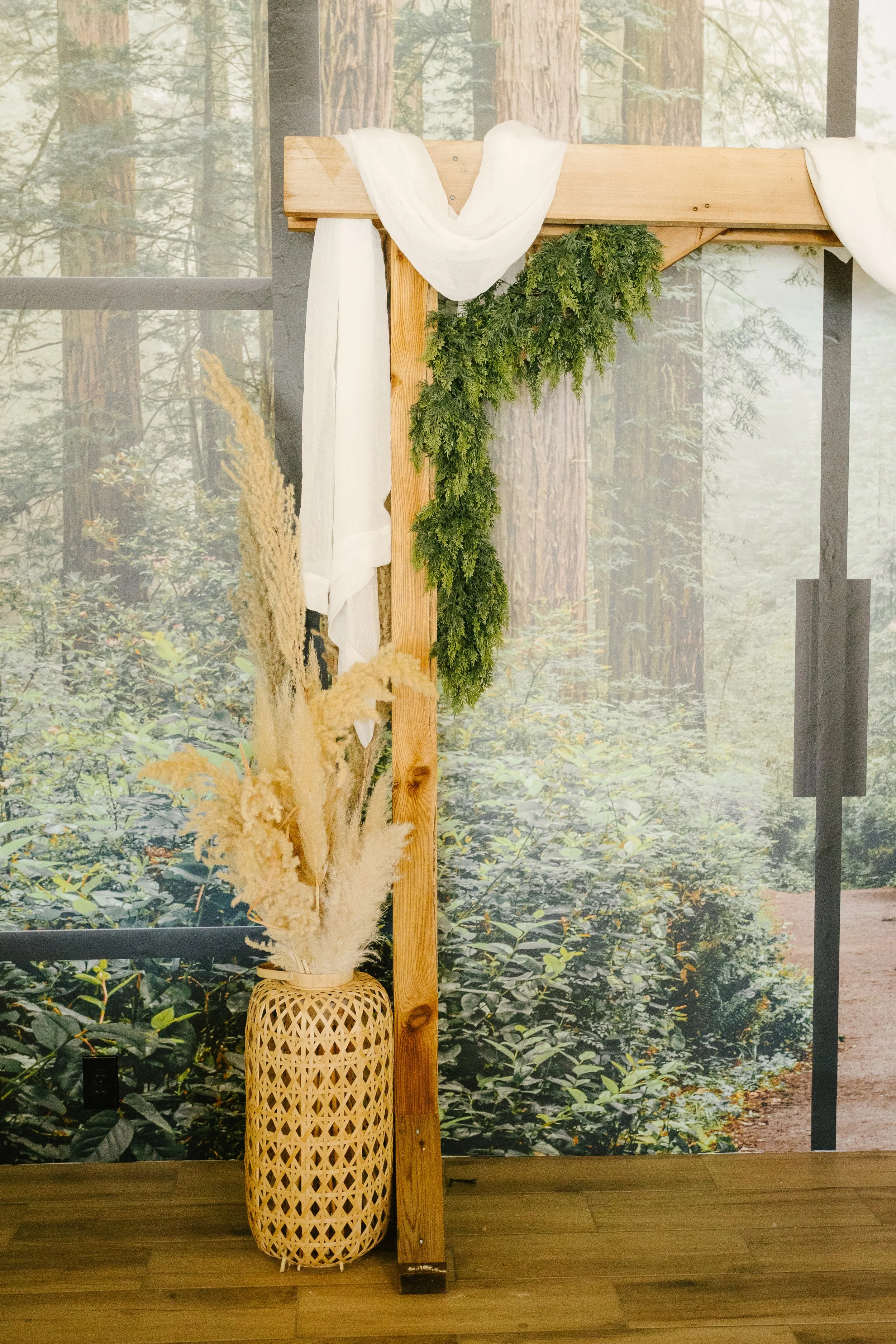 Decorative natural scene with a woven basket filled with pampas grass and greenery, positioned next to a wooden frame draped with white fabric, set against a forest-themed background.