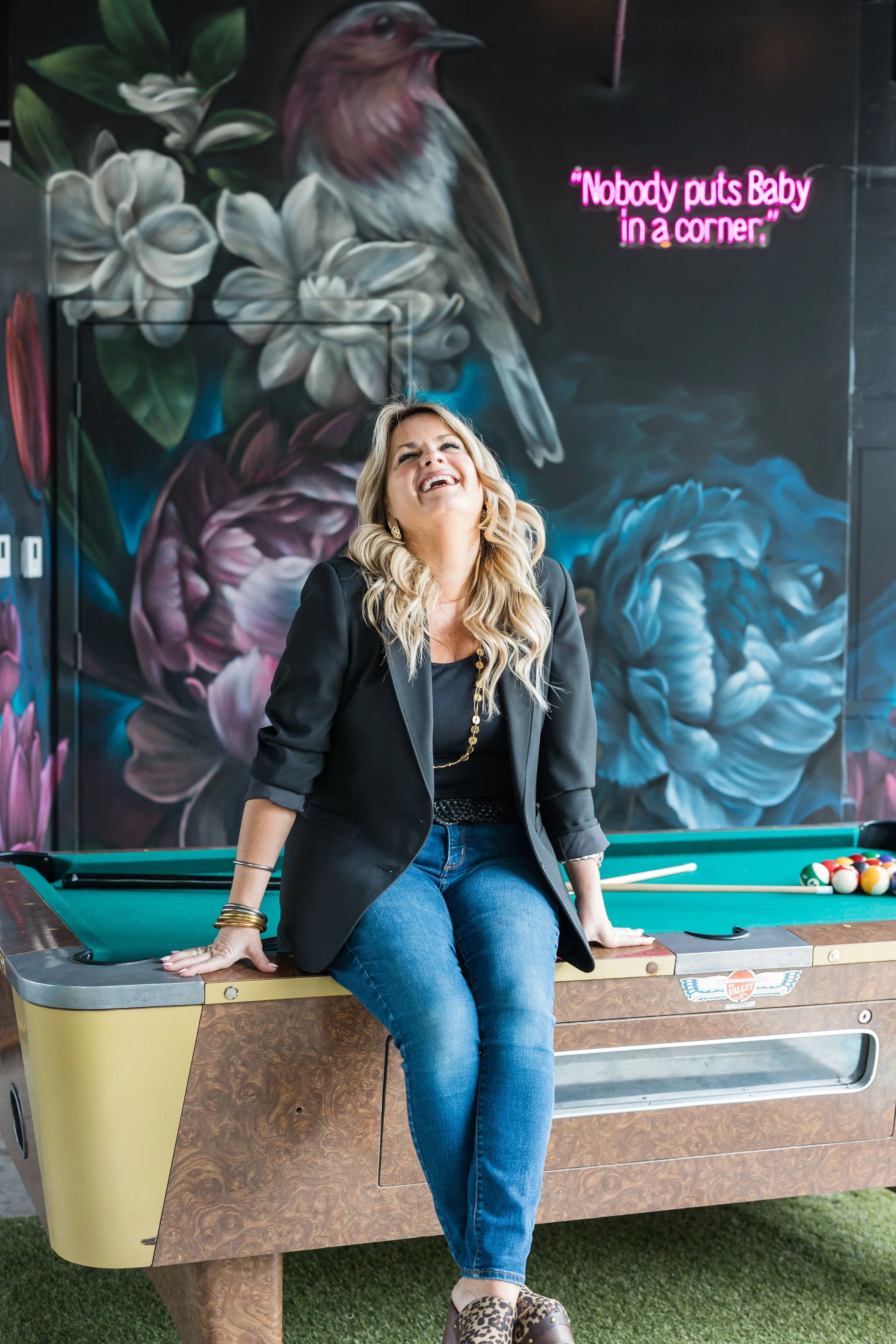 A woman with blonde hair wearing a black blazer, black top, and blue jeans sitting on a pool table, laughing and leaning back with one hand on the table. Behind her is a colorful mural of flowers and a bird, with a pink neon sign that reads 'Nobody puts Baby in a corner.'
