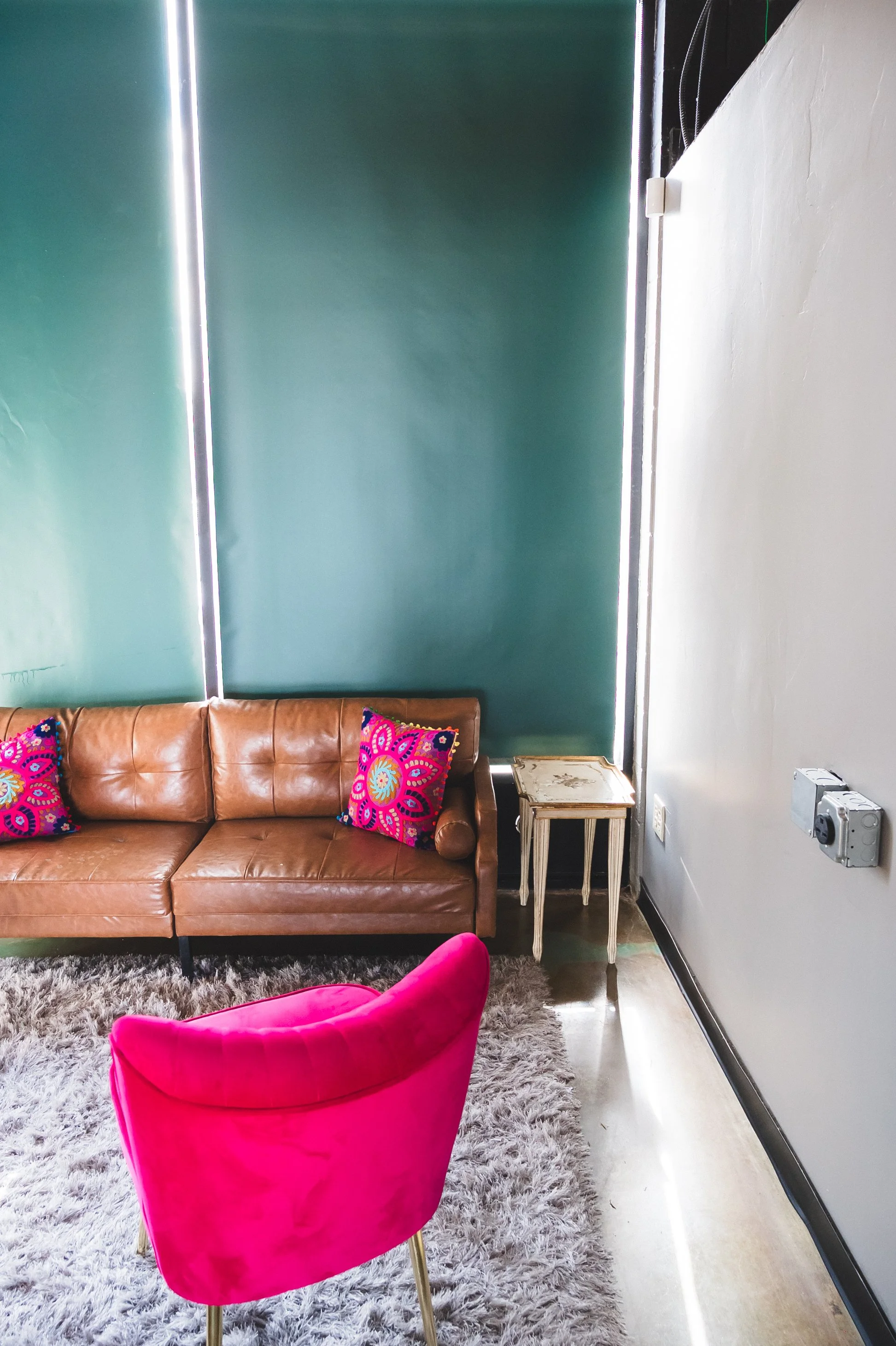A cozy living room corner with a brown leather couch, two colorful patterned pillows, a bright pink velvet armchair, a small ornate side table, a plush gray rug, and a teal accent wall with vertical light strips.