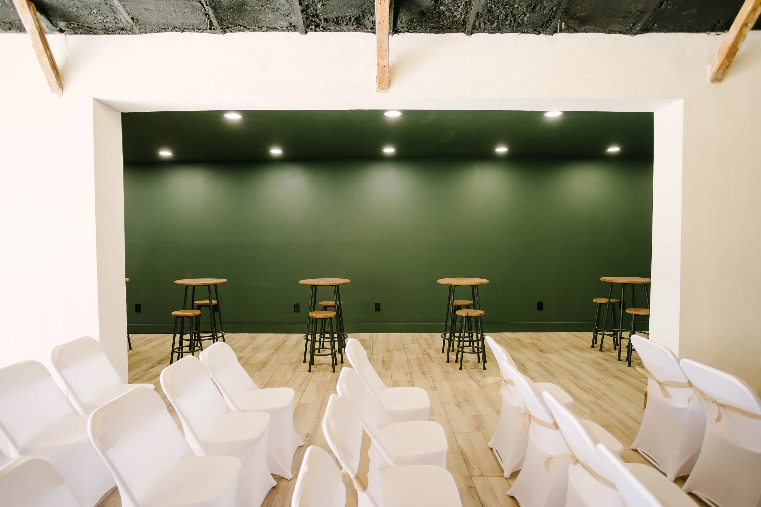 An indoor event space with white chairs covered in white fabric arranged in rows facing a green accent wall with six ceiling spotlights. There are four tall tables with wooden tops and black metal legs positioned along the green wall.