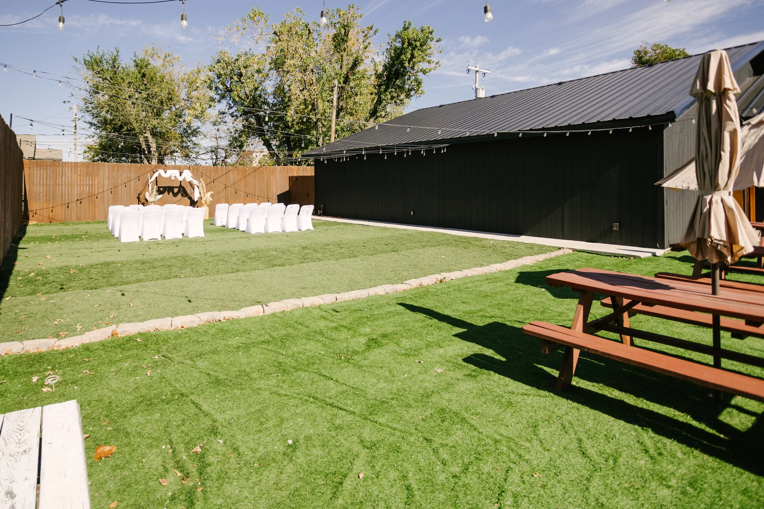 Outdoor backyard decorated for an event with white chair covers, a decorated arch, string lights, and a picnic table with an umbrella.