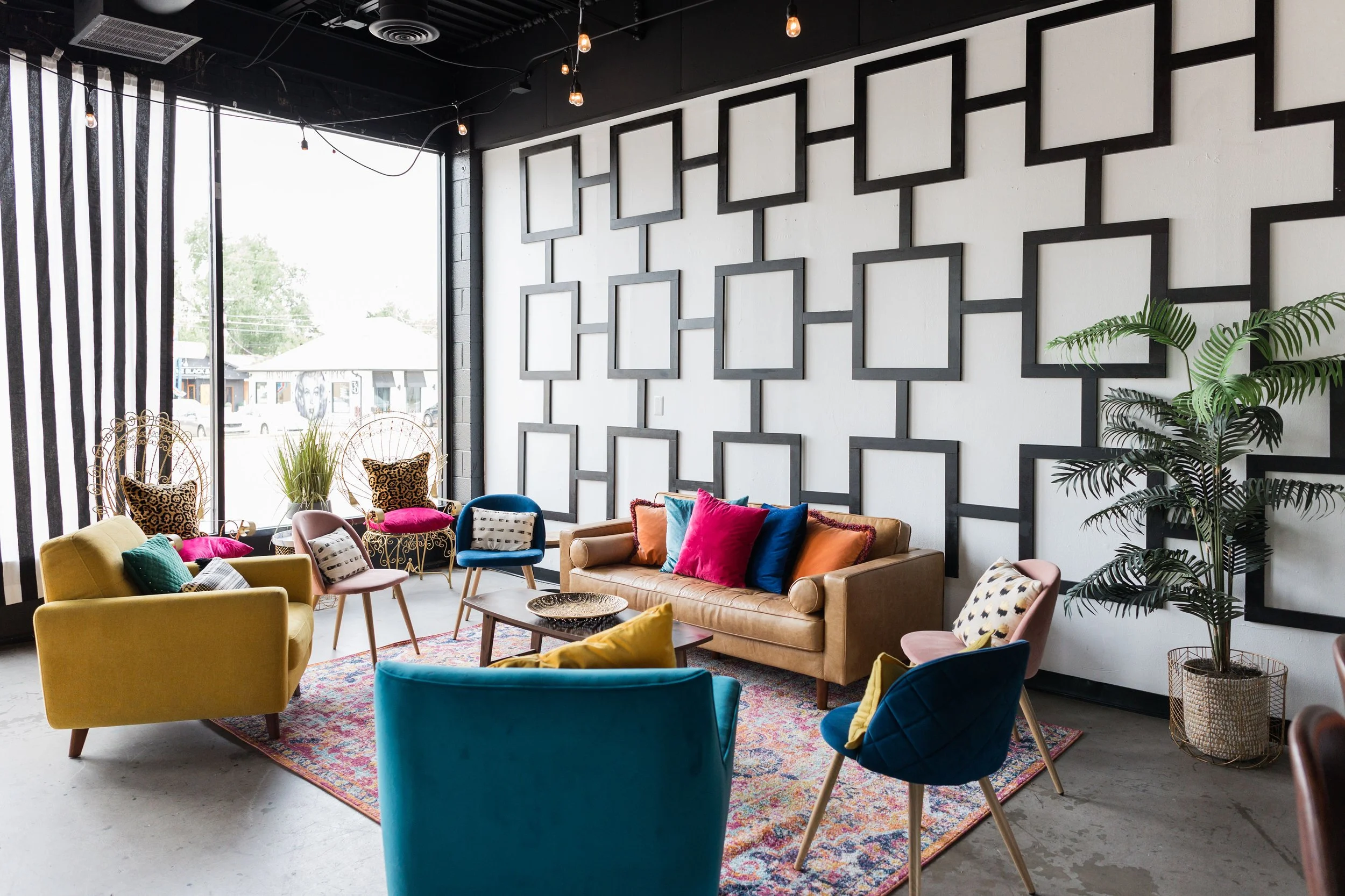 Modern living room with colorful sofa, armchairs, decorative pillows, and a patterned rug, near large windows with striped curtains, and framed wall art.