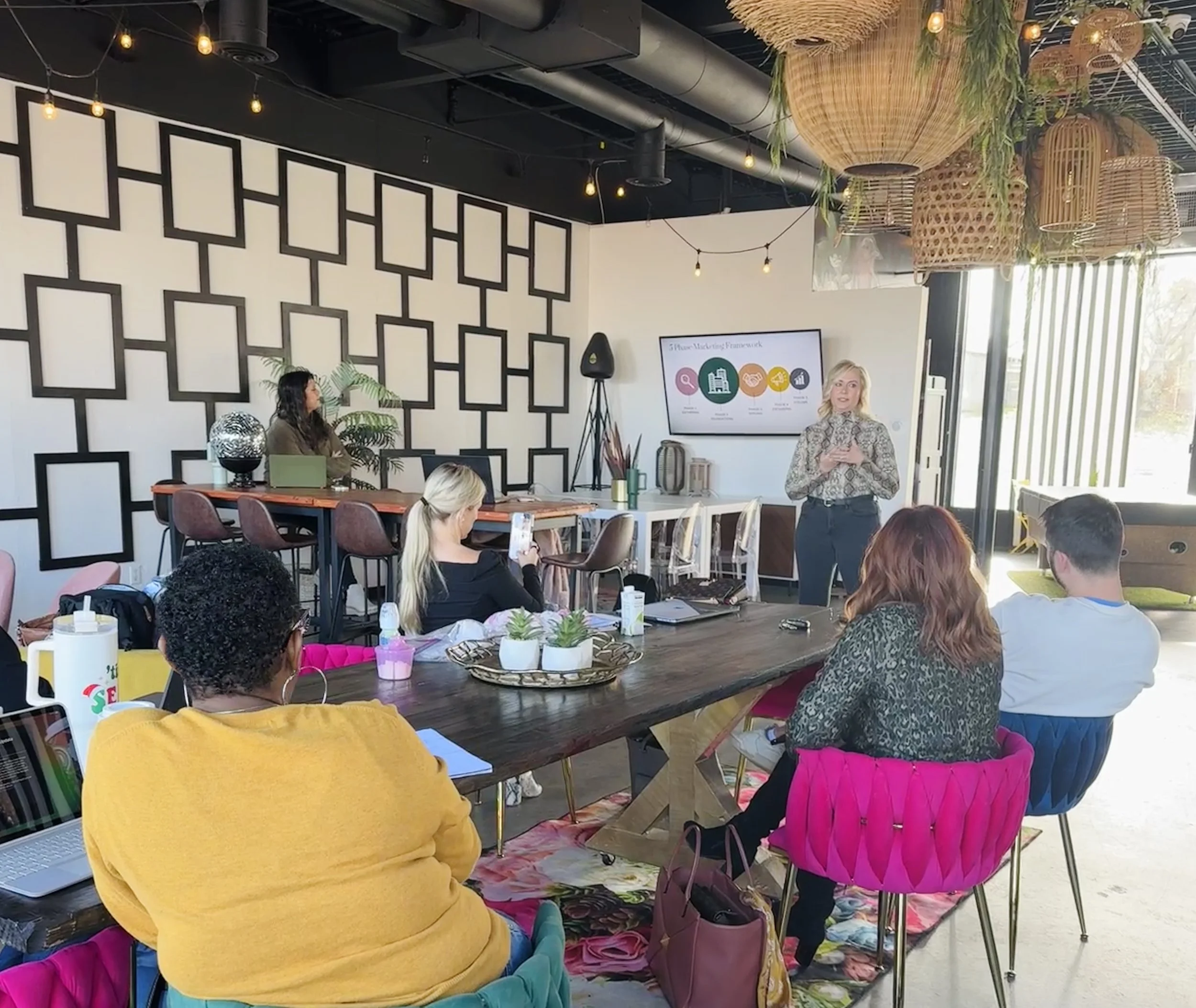 A woman presenting in a modern, well-lit room to a small audience seated around a wooden table, with a large screen displaying a presentation behind her.
