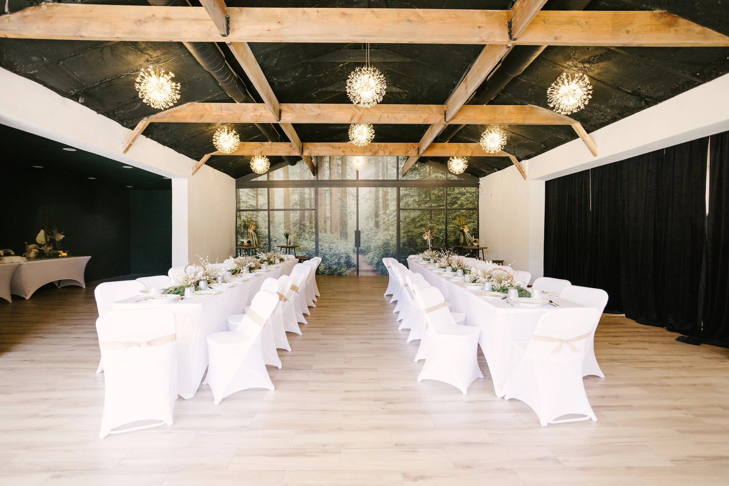 Elegant event space with two long white-draped tables set with centerpieces, surrounded by white chairs with beige sashes. The room has a high ceiling with exposed wooden beams and spherical light fixtures, with a large forest mural on the back wall.