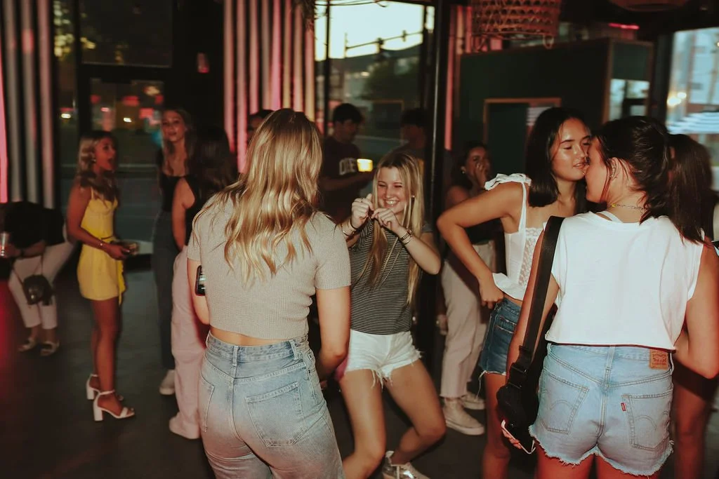 Group of young women dancing and socializing in a dimly lit indoor space, possibly a bar or club, with some holding drinks and others engaged in conversation.