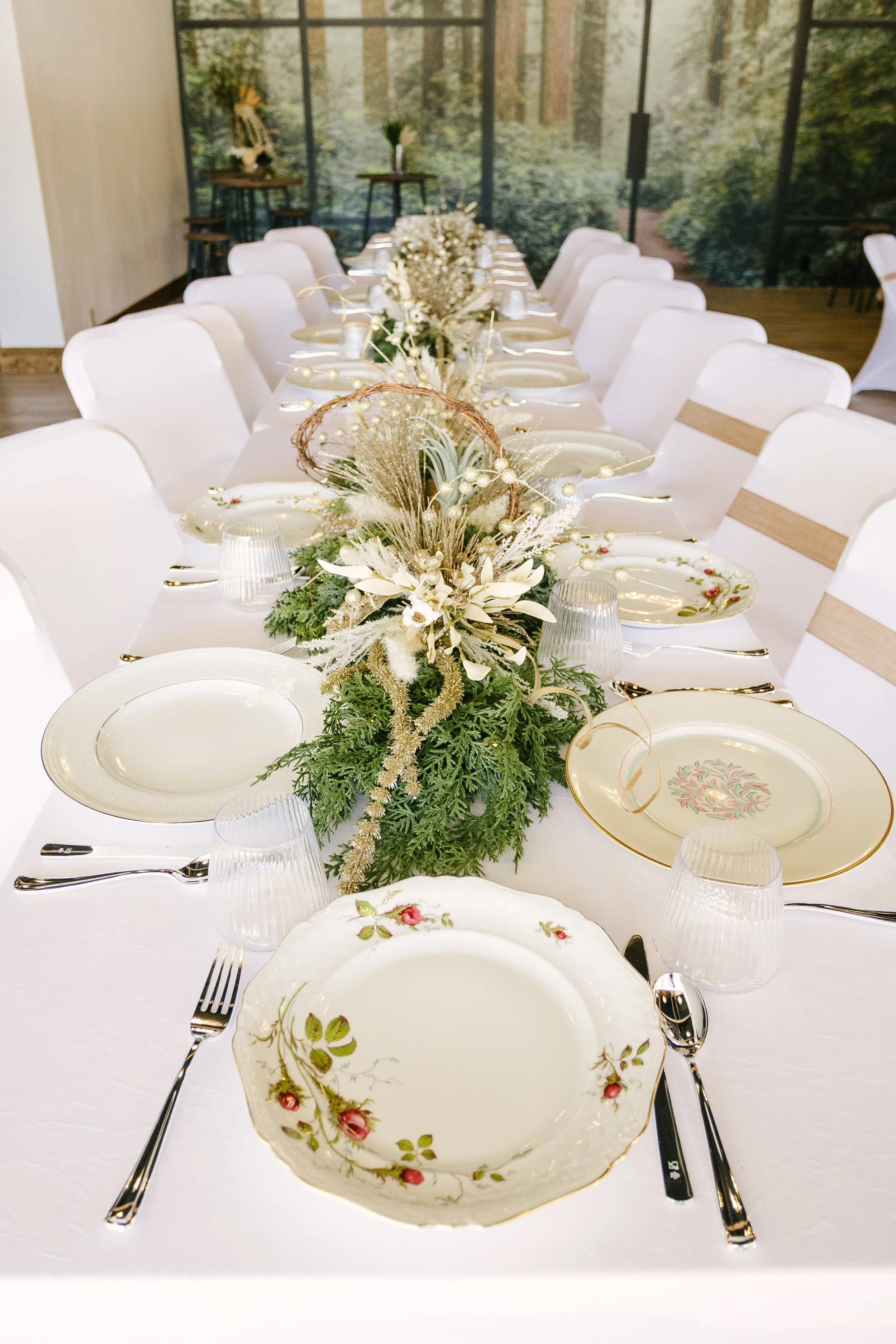 Decorated dining table set for a special occasion with floral arrangement centerpiece, vintage-style plates, silverware, and glassware, in a room with a nature-themed mural.