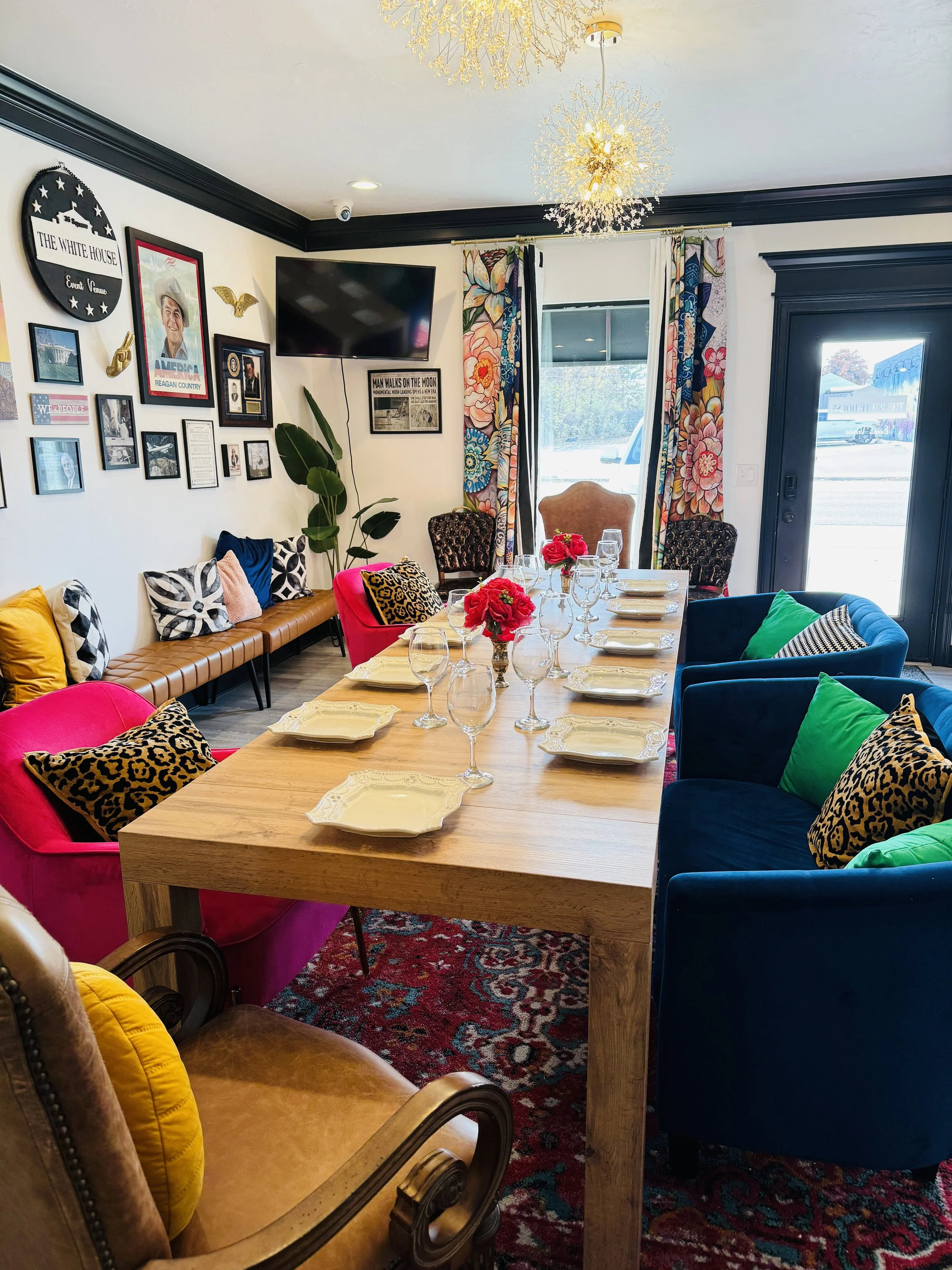 A dining room with a wooden table set for eight, decorated with pink flowers in the center and glassware. The room has colorful upholstered chairs, patterned pillows, and a gallery wall featuring artwork, flags, and memorabilia. There are large curta