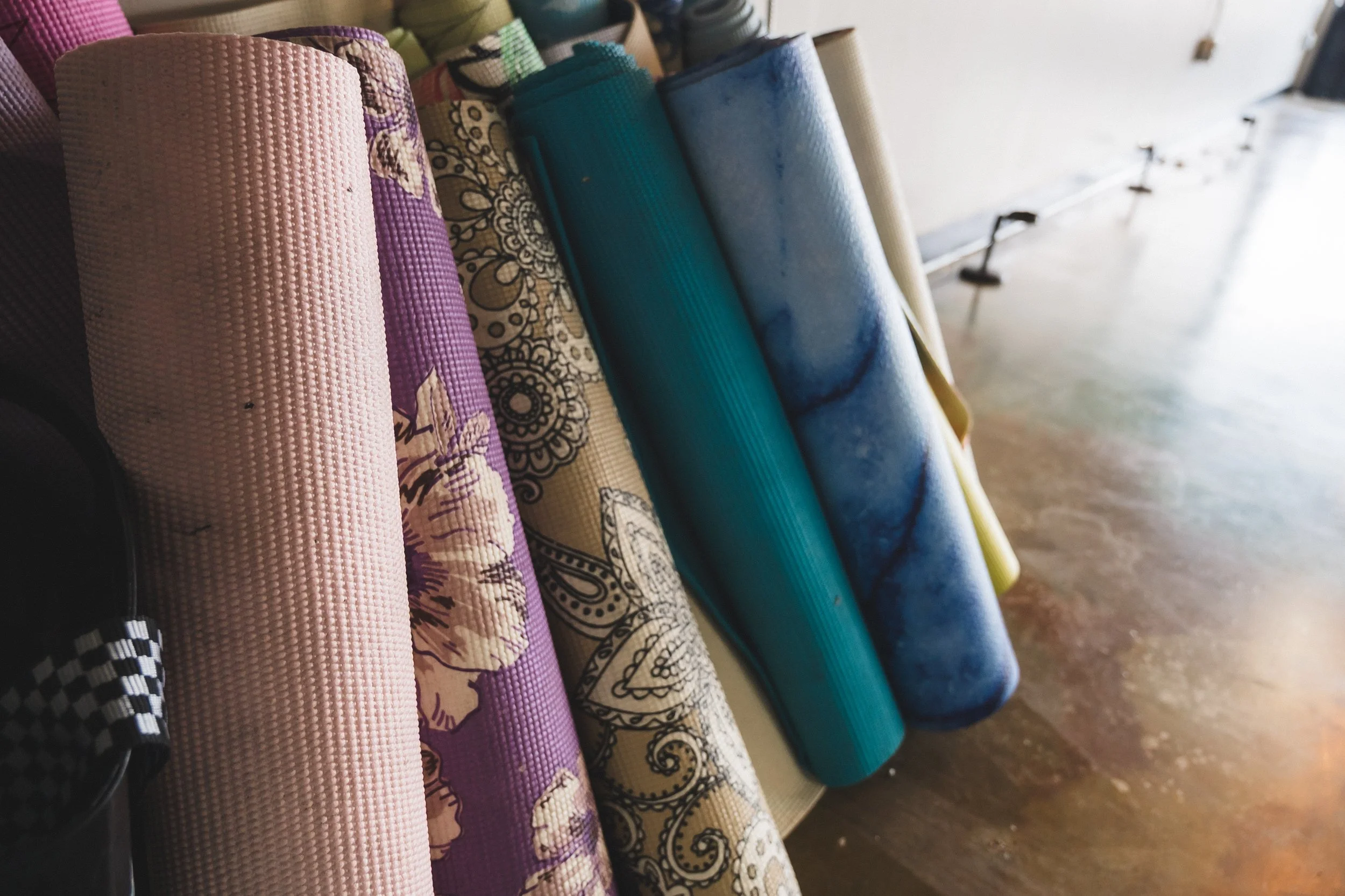 Assorted rolled yoga mats with patterns and solid colors leaning against a wall inside a building.