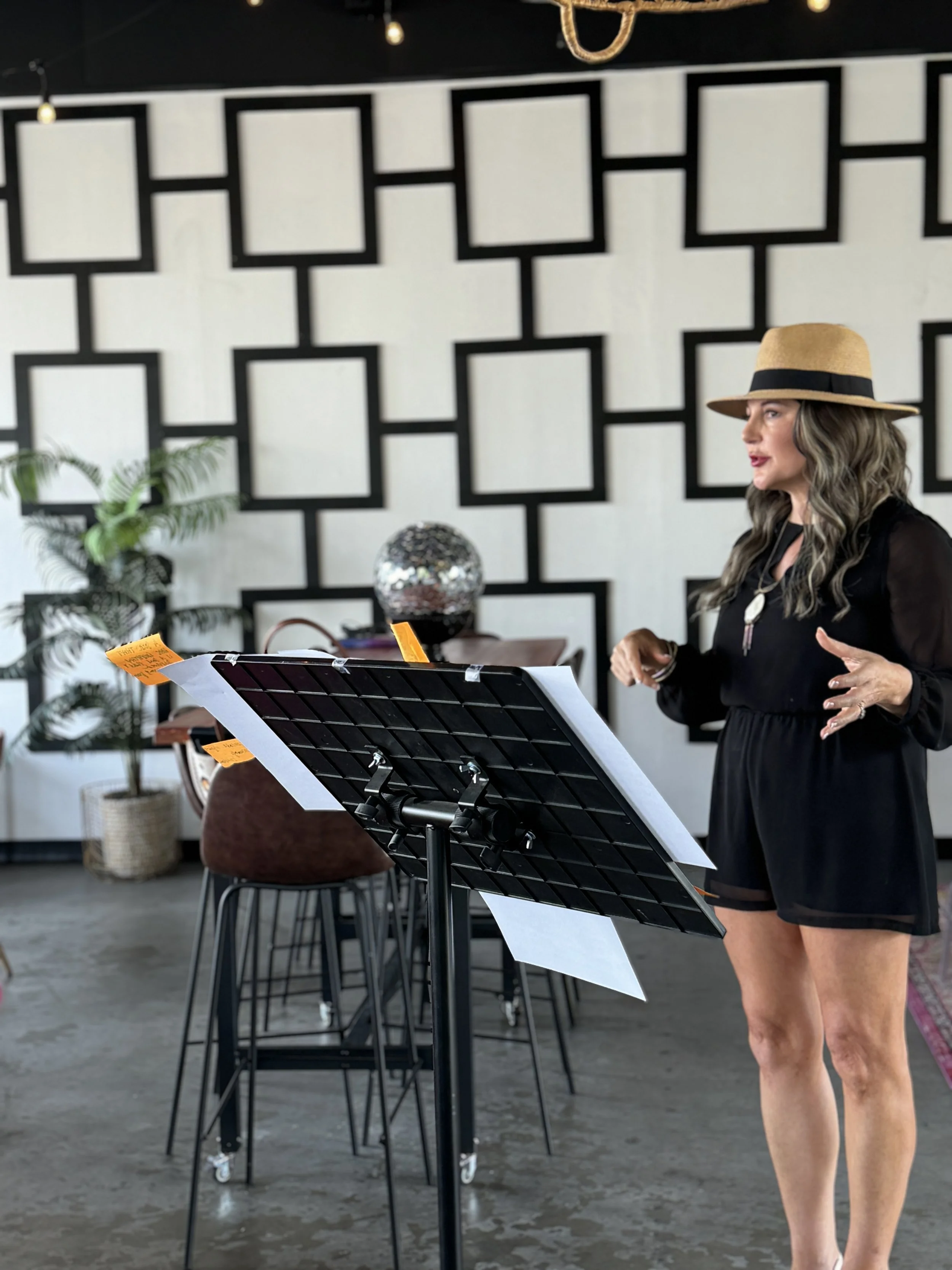 A woman with wavy hair wearing a wide-brimmed hat and black outfit standing and talking in front of a music stand with sheet music, in a decorated indoor space with geometric wall art, a potted plant, and bar stools.