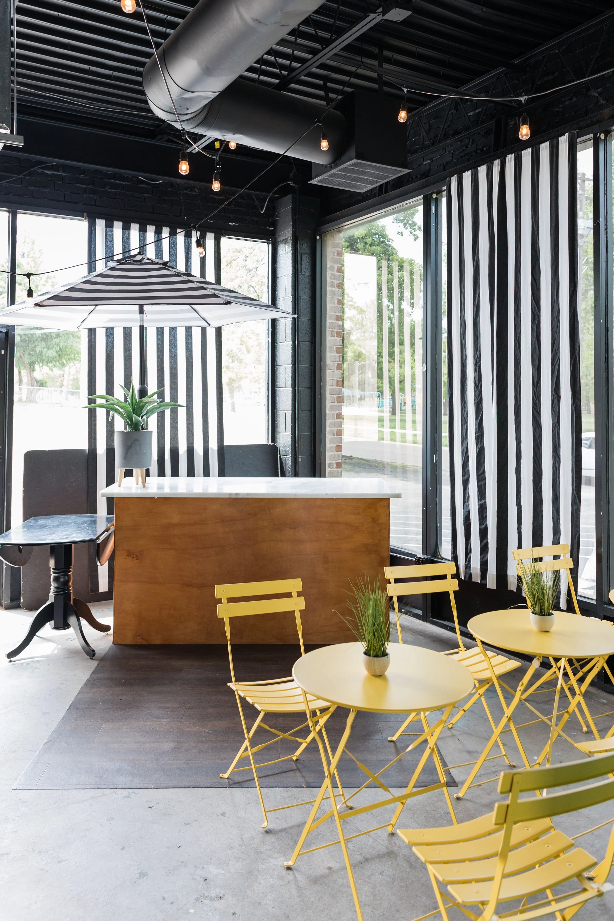 Bright coffee shop interior with yellow tables and chairs, black and white striped curtains, a large window, and an umbrella with a plant on a counter.