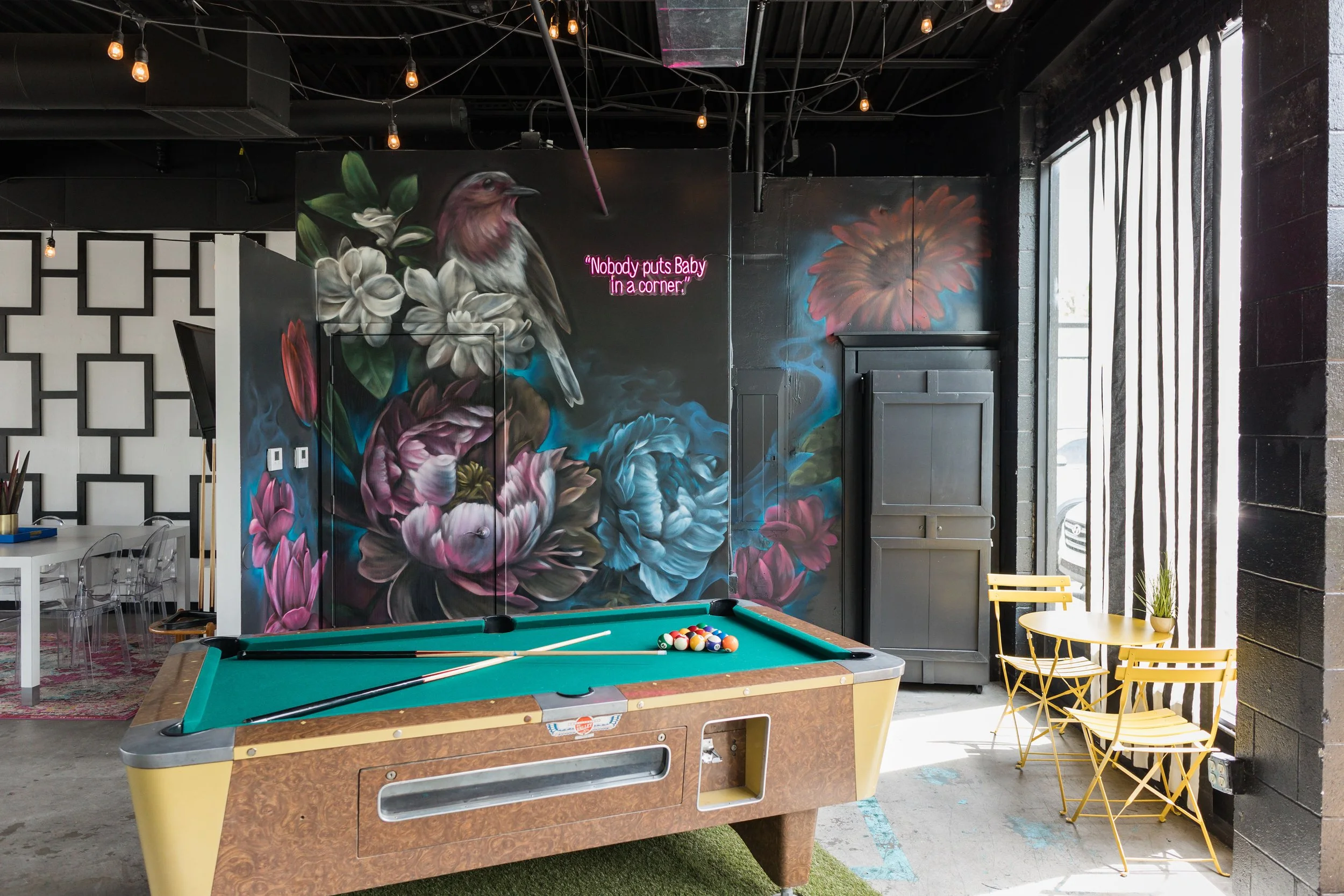 Indoor scene with a pool table, floral wall mural, neon sign, yellow chairs, and window with vertical blinds.