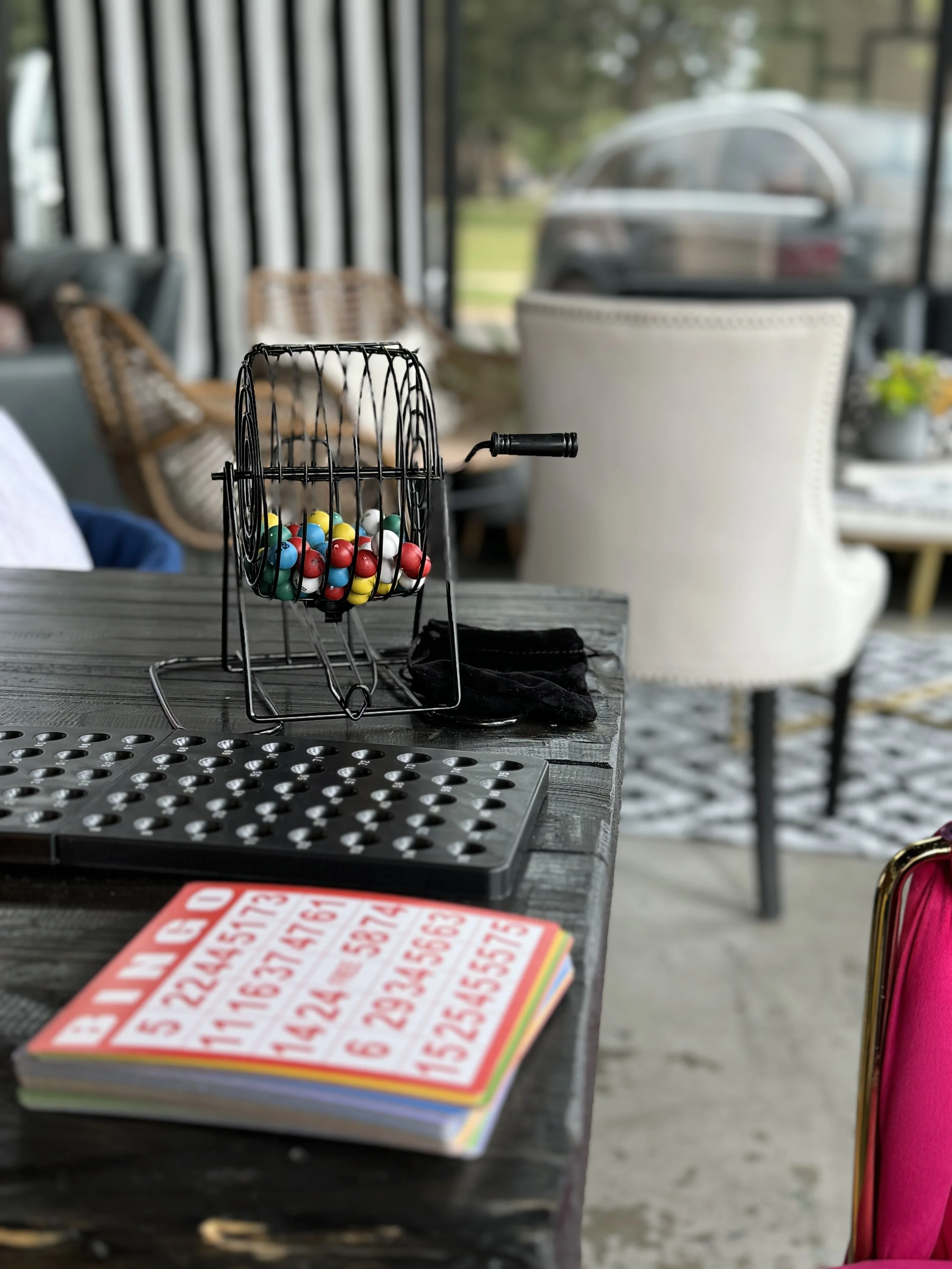 Bingo game setup on a table with a cage full of colorful bingo balls, a bingo number card, and a black drawstring bag inside a cozy room with chairs and large windows.