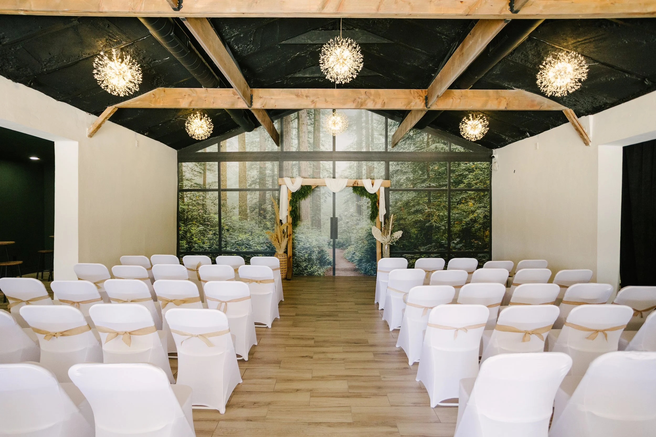 Indoor wedding ceremony setup with white chairs, a decorated arch, and forest-themed backdrop.
