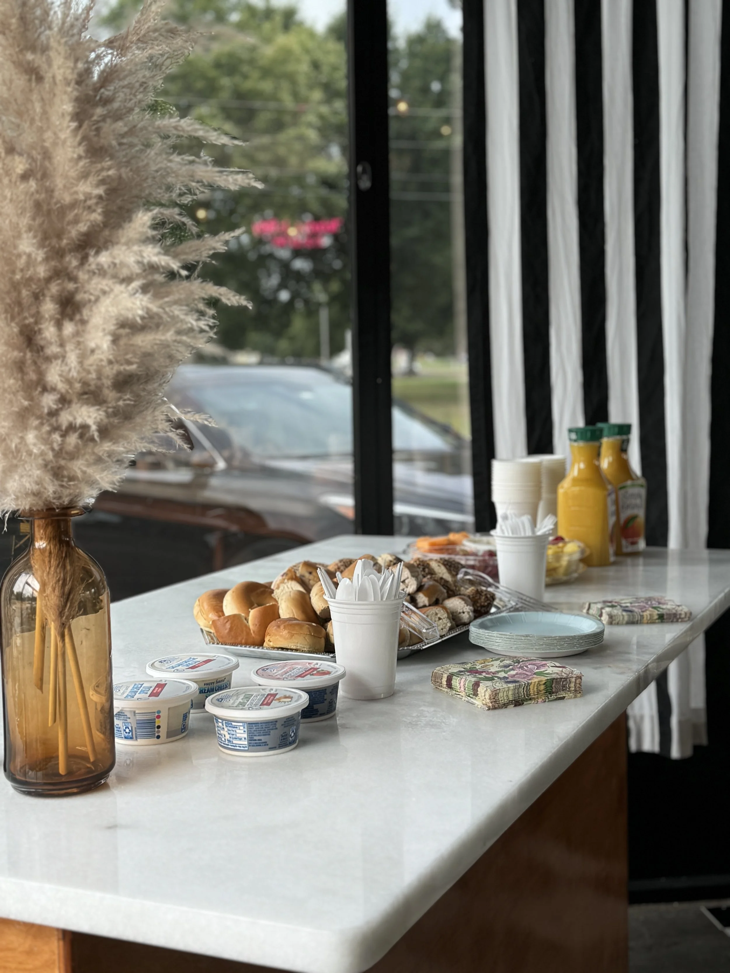 A breakfast buffet on a table with pastries, yogurt, condiments, plates, napkins, and utensils inside a restaurant with window views of greenery and parked cars outside.