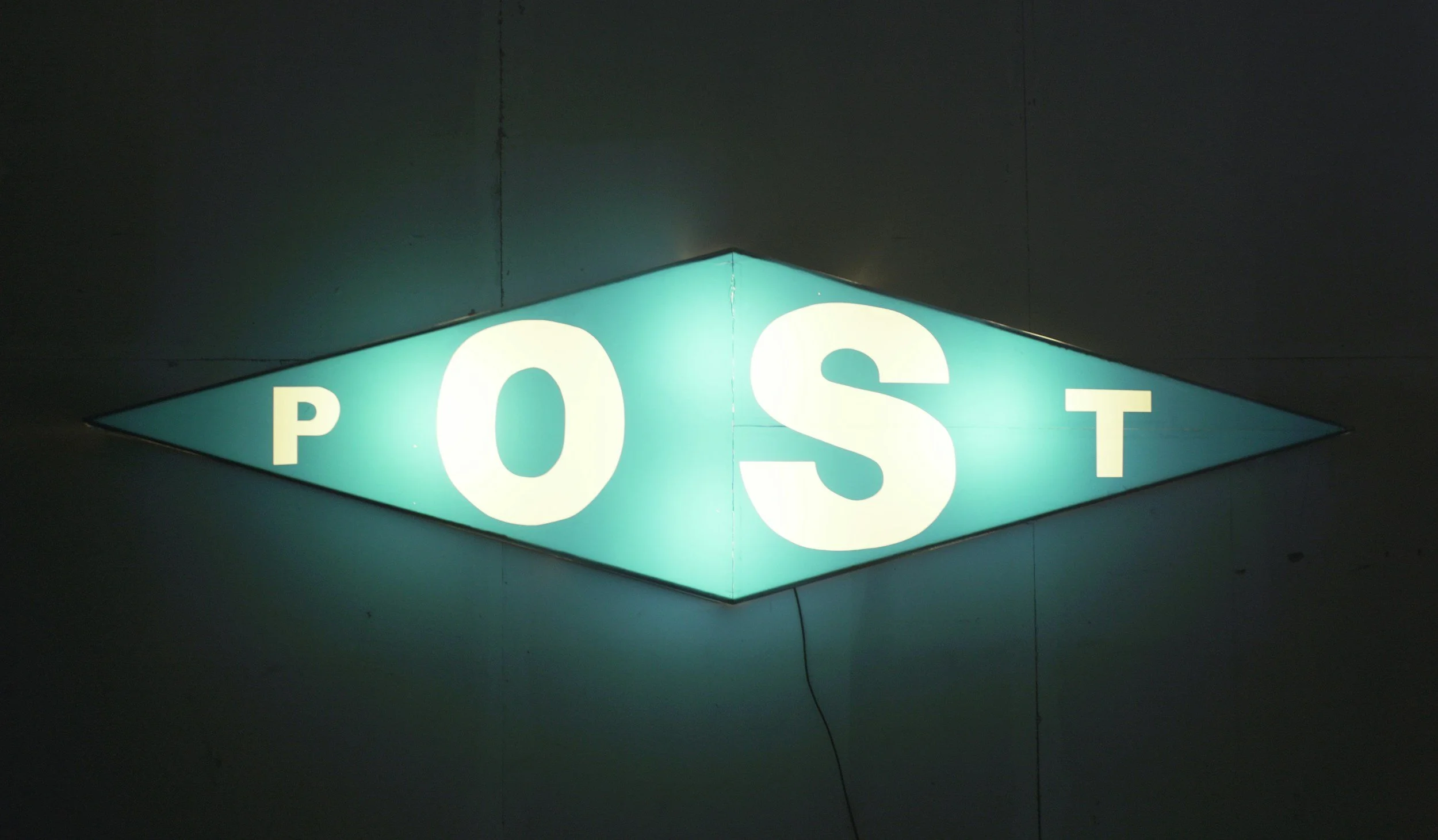 Illuminated sign with the word 'POST' in large white letters on a teal background, shaped like a diamond.