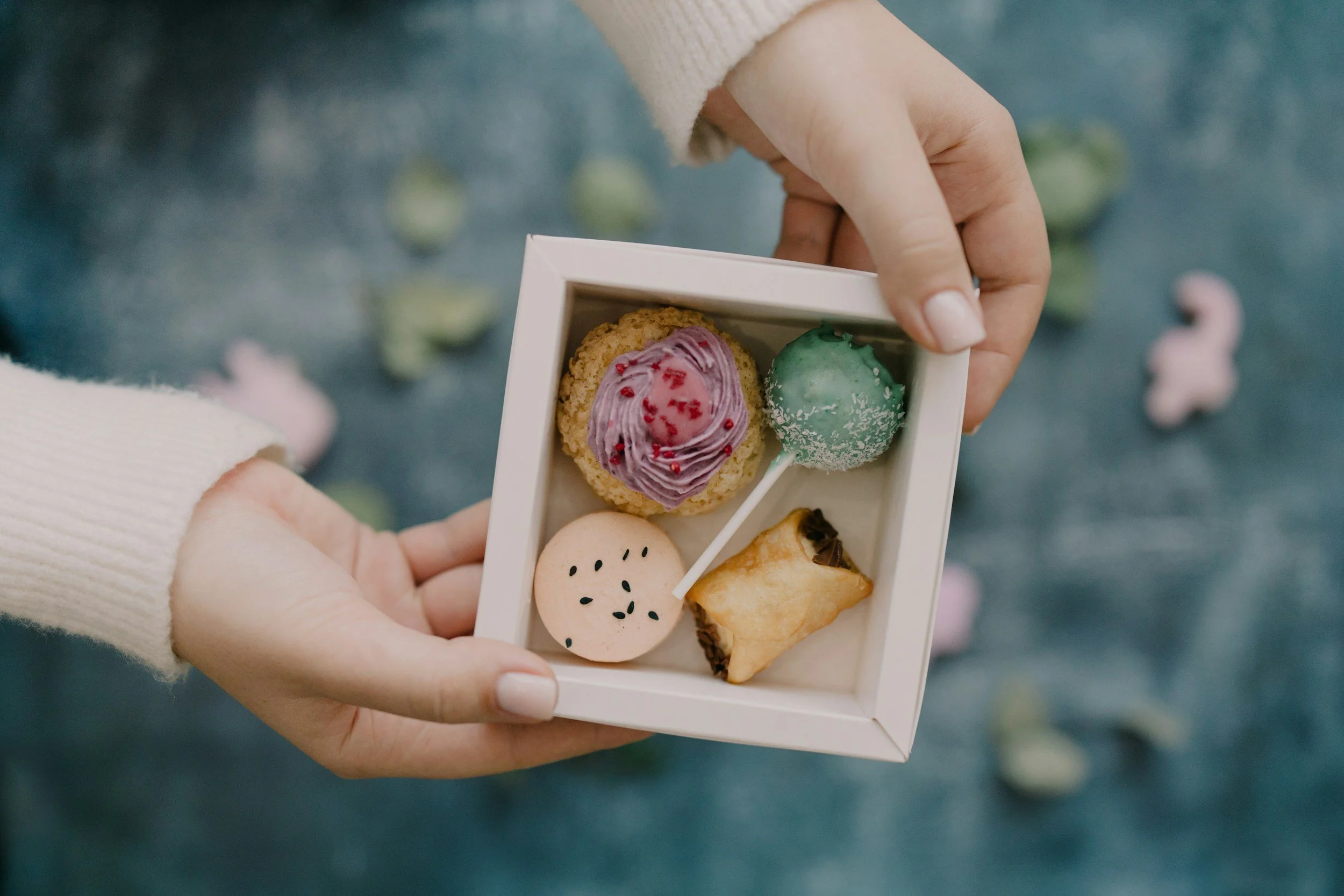 Person holding a white box containing a cookie, a green cake pop, a pink macaron, and a pastry. Background has scattered pastel-colored bunny and chick-shaped candies.