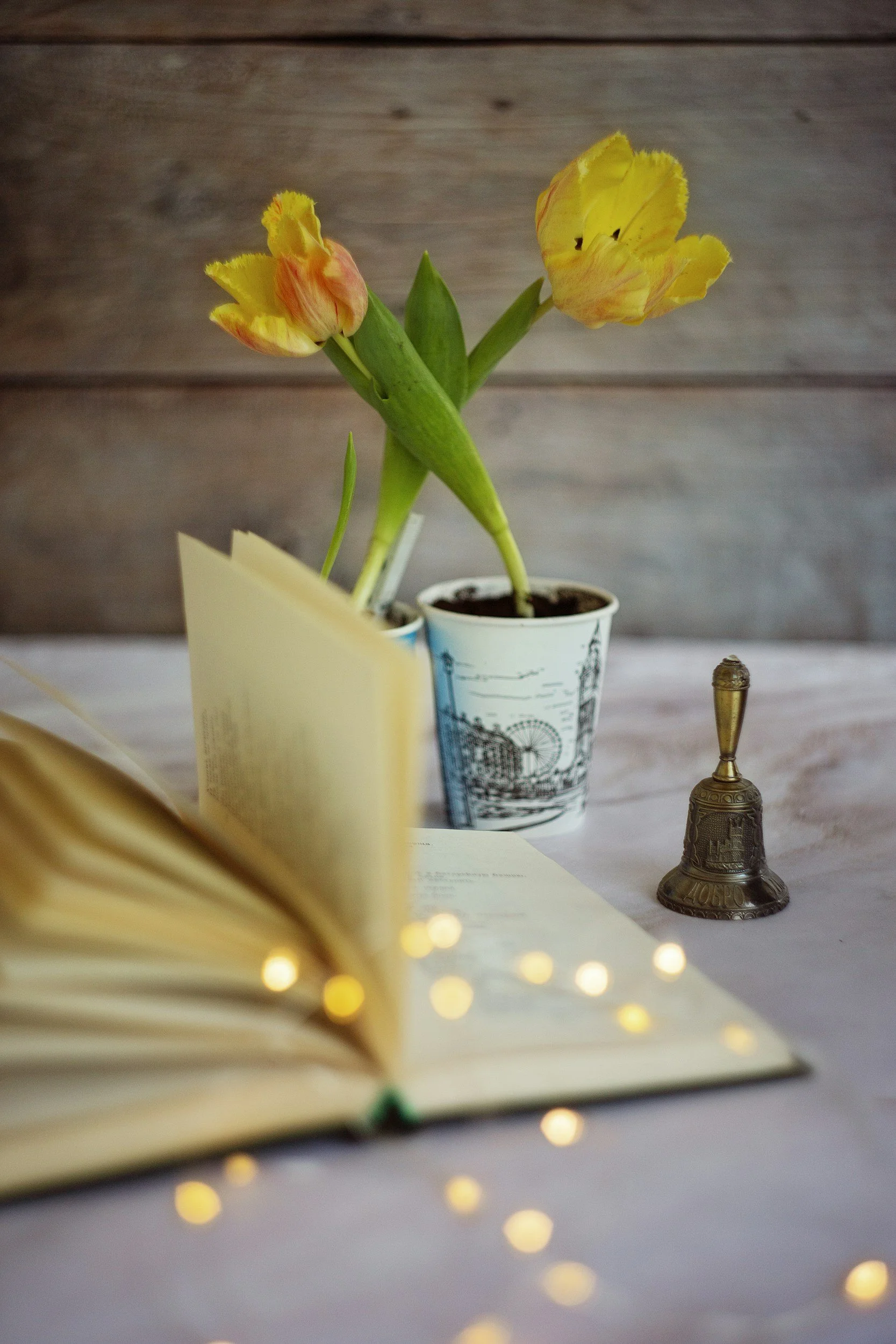 Yellow tulips in a decorative paper cup, an open book with yellow fairy lights in front, and a small brass bell on a white surface with a wooden wall background.