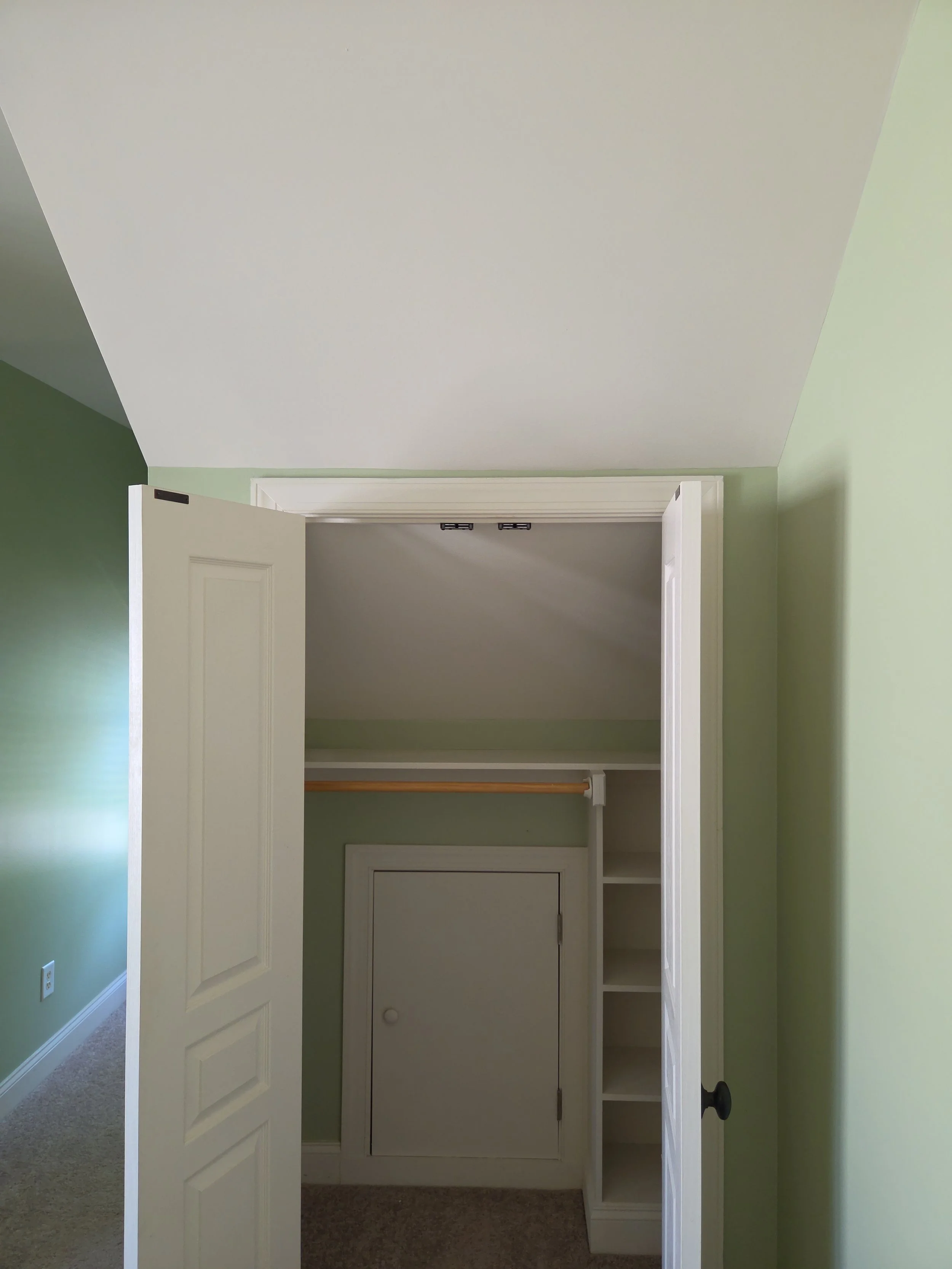 Empty closet space with an open door, painted green walls, and built-in shelves, ceiling, and a small door at the back of the closet.