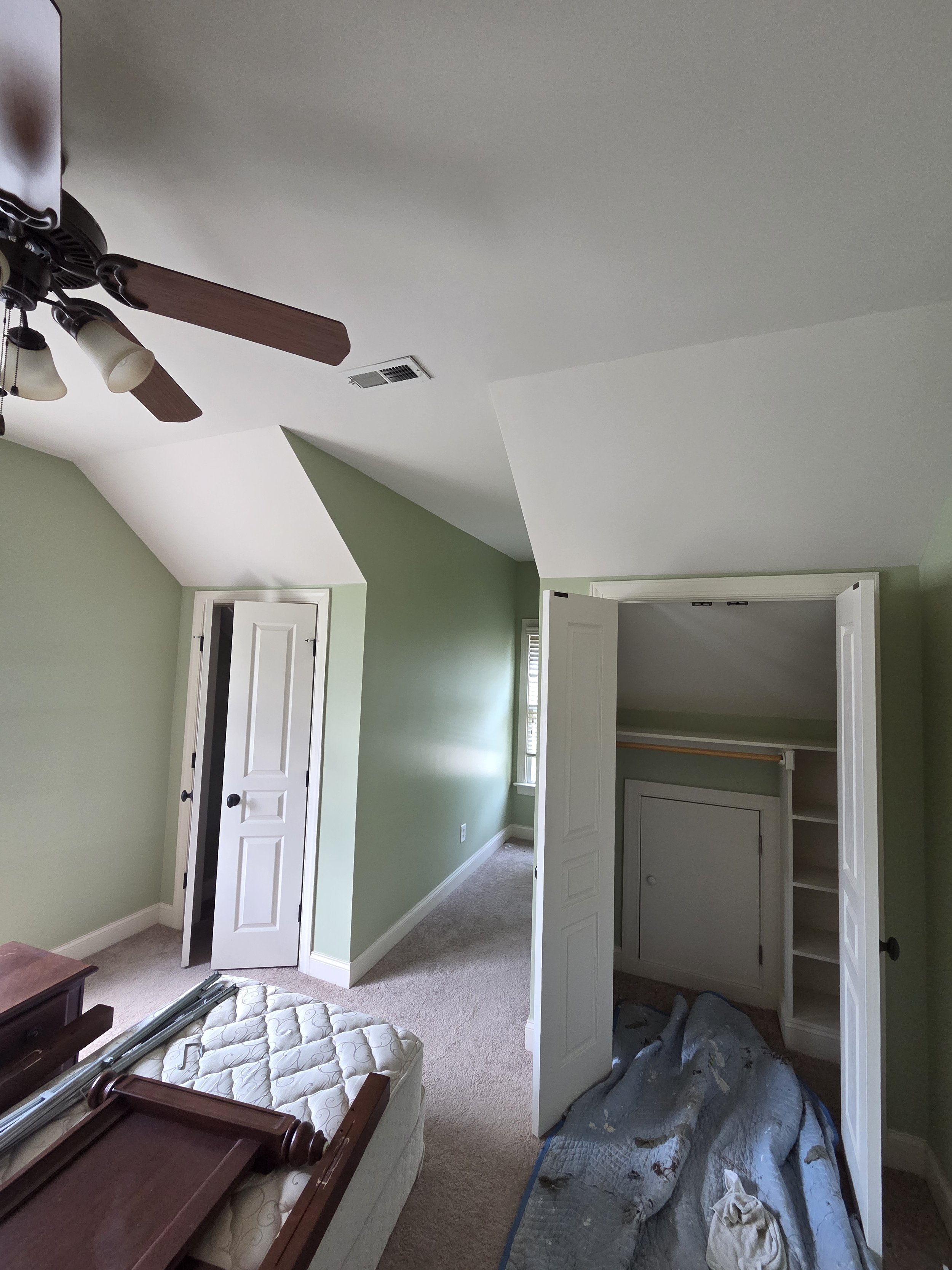 A room under renovation with an open closet, a ceiling fan with lights, a small window, a closed door, and a partially disassembled bed frame and mattress.