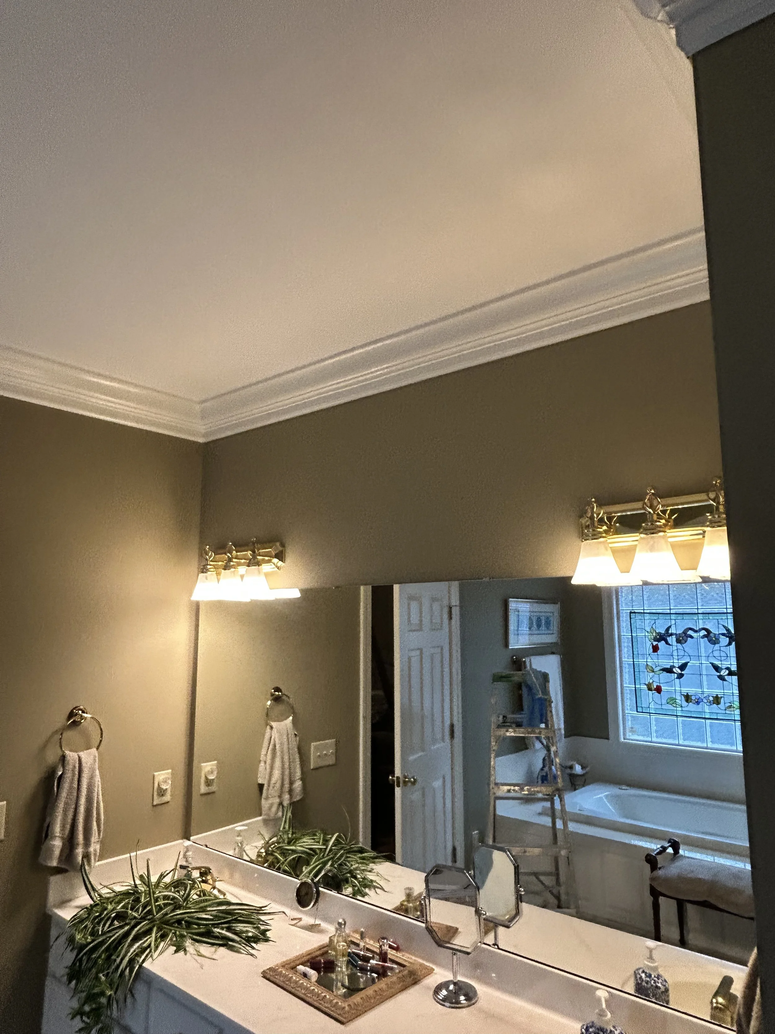 Bathroom vanity mirror reflecting a bathroom with a window, decorative stained glass, ladder, and a bathtub, with walls painted in a dark beige color.