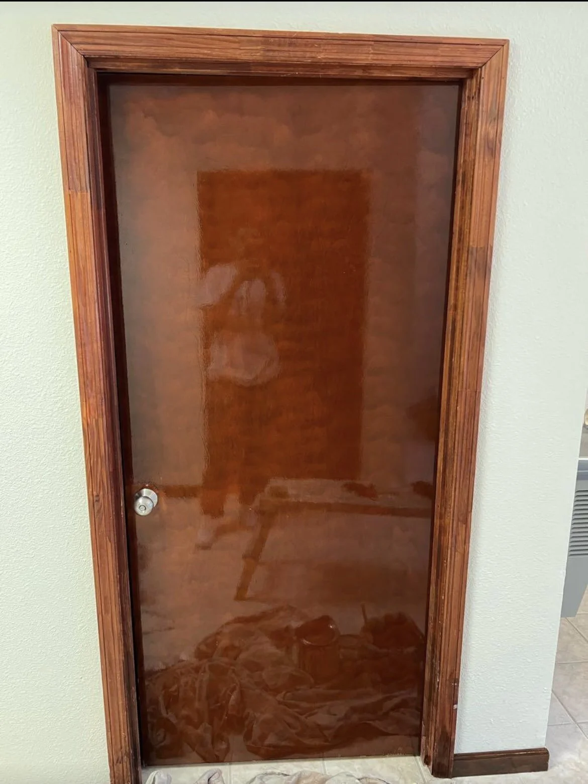 A wooden framed glass door with a reflective surface, showing a person's reflection and some objects on the floor, against a beige wall.