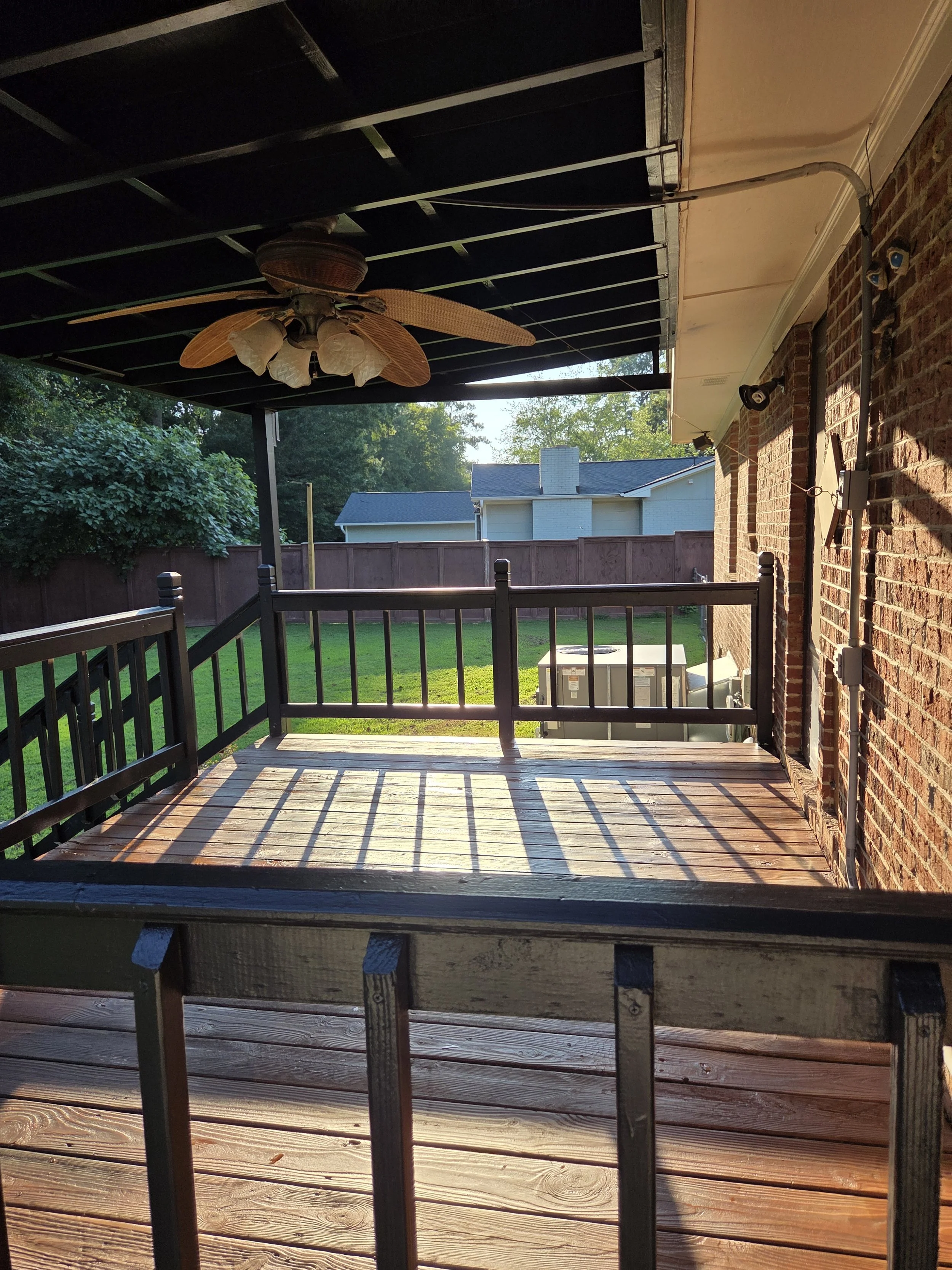 Backyard patio with wooden deck, black metal railing, ceiling fan with wicker blades, brick house wall, outdoor lighting, electrical wiring, green grass, and neighboring houses in the background.