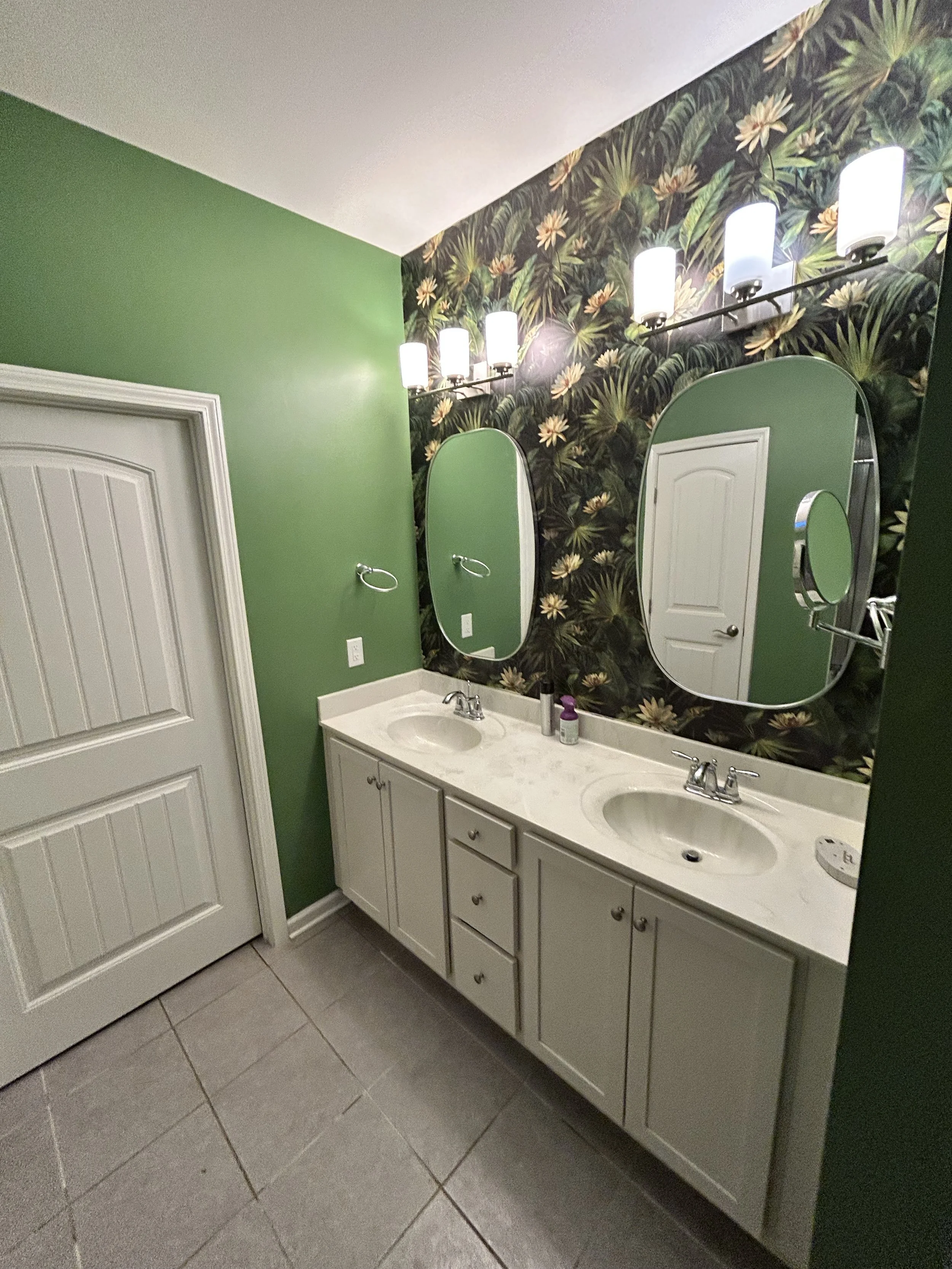 Double bathroom vanity with two sinks, oval mirrors, green wall behind, floral wallpaper on the upper half, and a light fixture with six white lights above the mirrors.