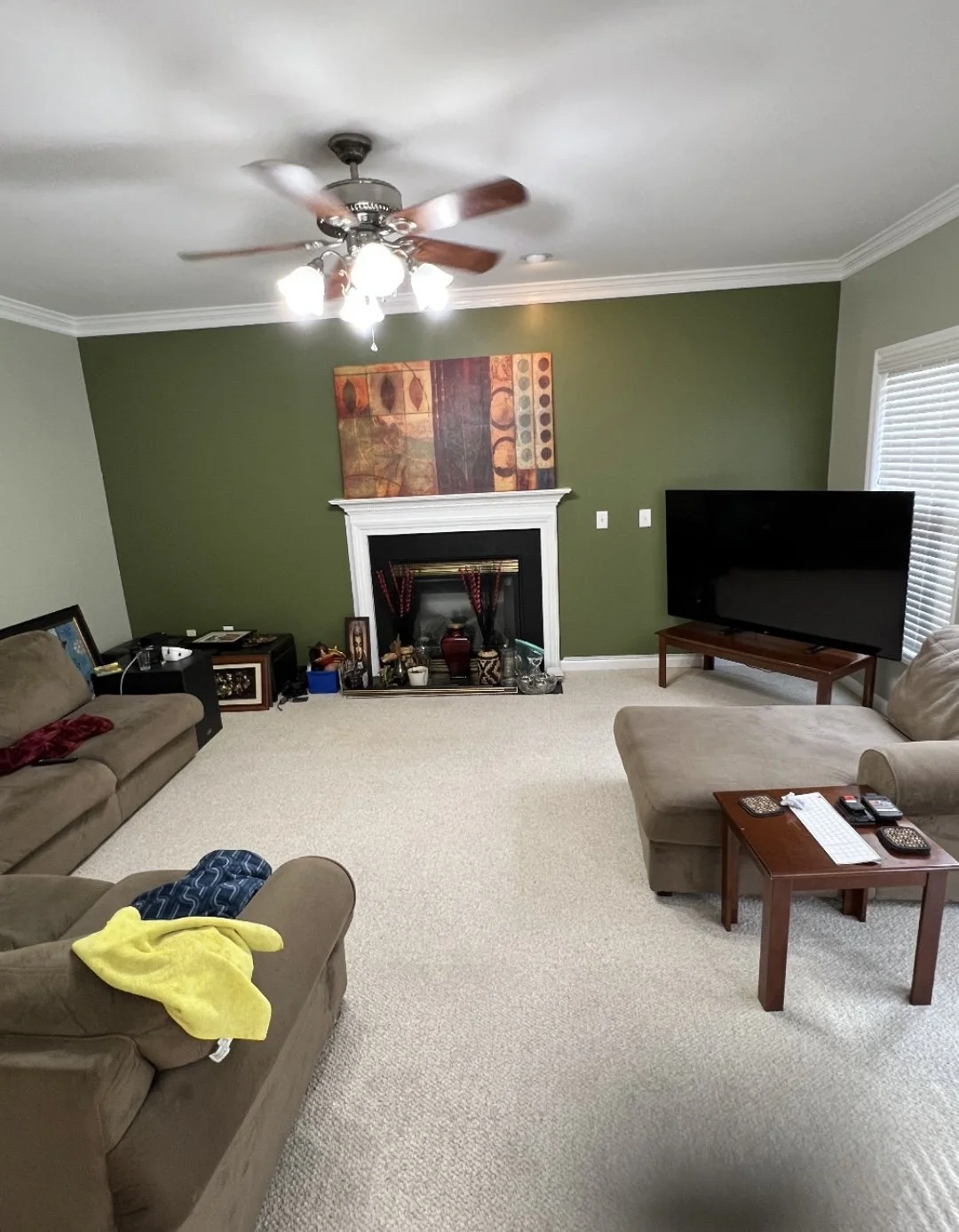 Living room with a green accent wall, carpeted floor, tan couches, a flat-screen TV on a stand, a coffee table with remotes and papers, a fireplace with decorative items, and a ceiling fan with lights.